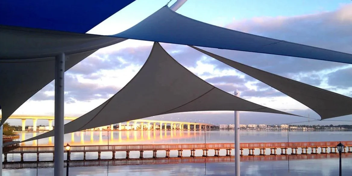 View of a city skyline across a river, with modern shade sails and a wooden pier in the foreground, and a bridge in the background during cloudy weather. Commercial shade sails. commercial shade awning, shade canopy, shade canopies.