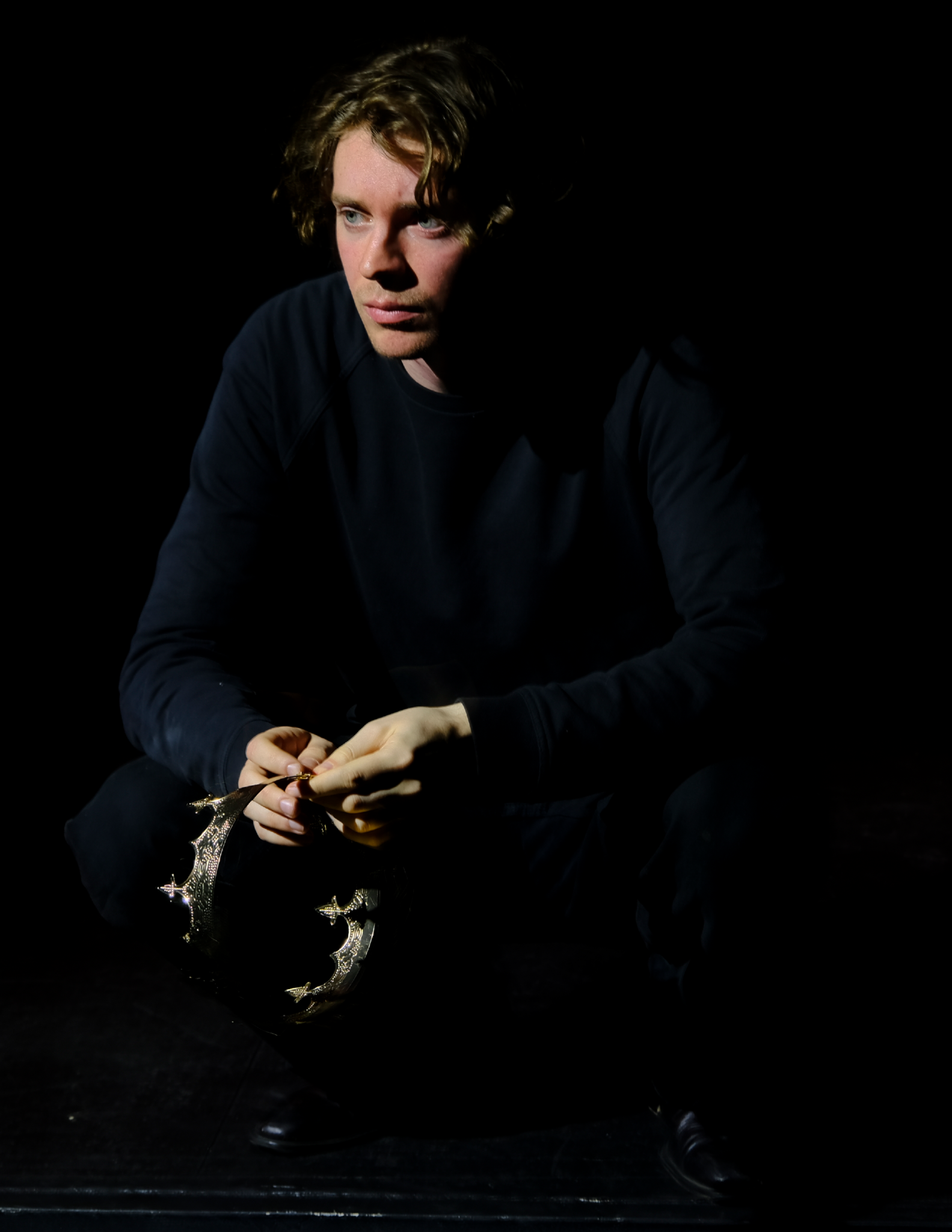 A young man with wavy blonde hair, dressed in black, crouching in darkness while holding a decorative crown.