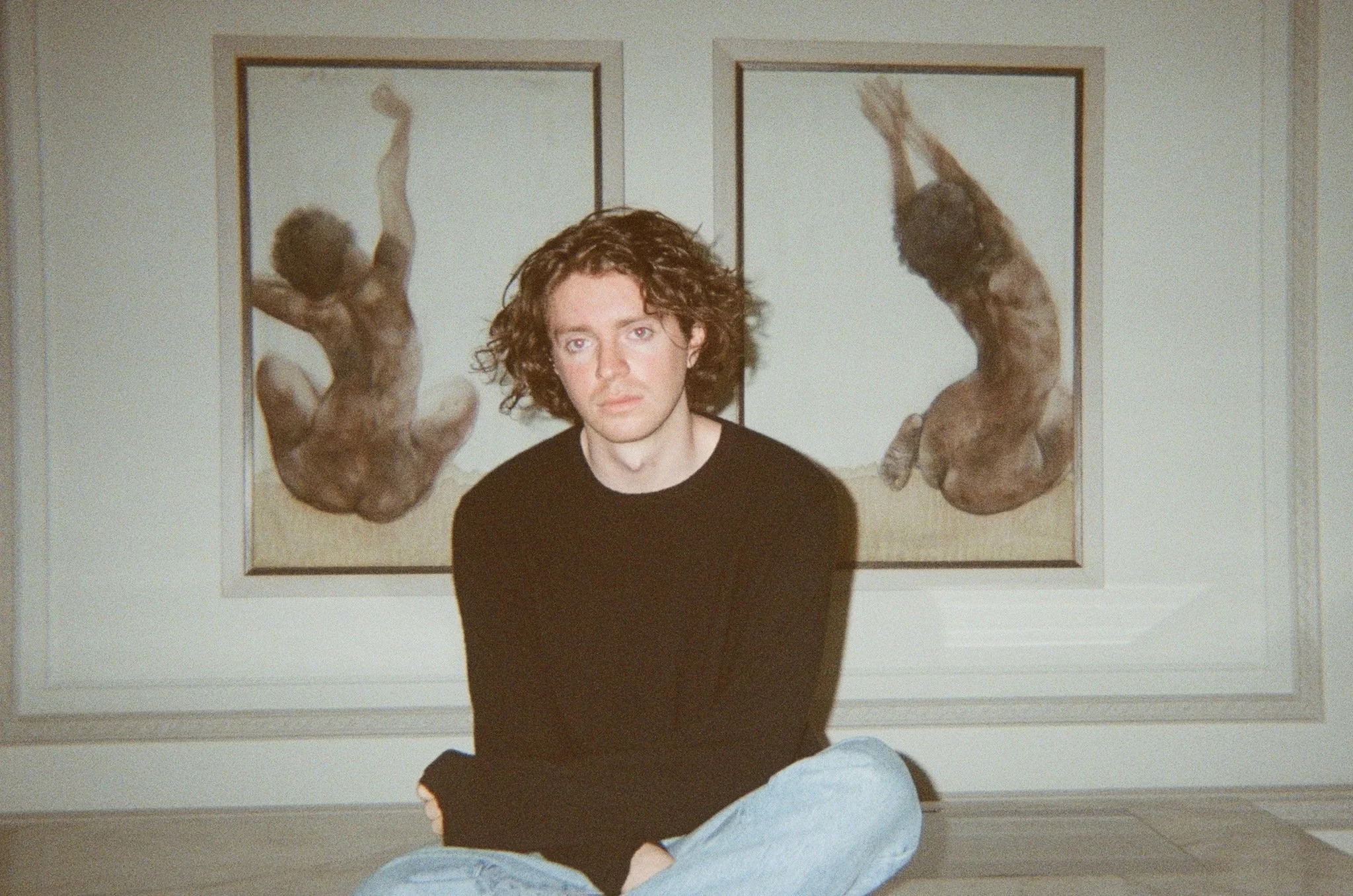 Close-up portrait of a young man with medium-length curly brown hair, blue eyes, and light skin, wearing a dark shirt.