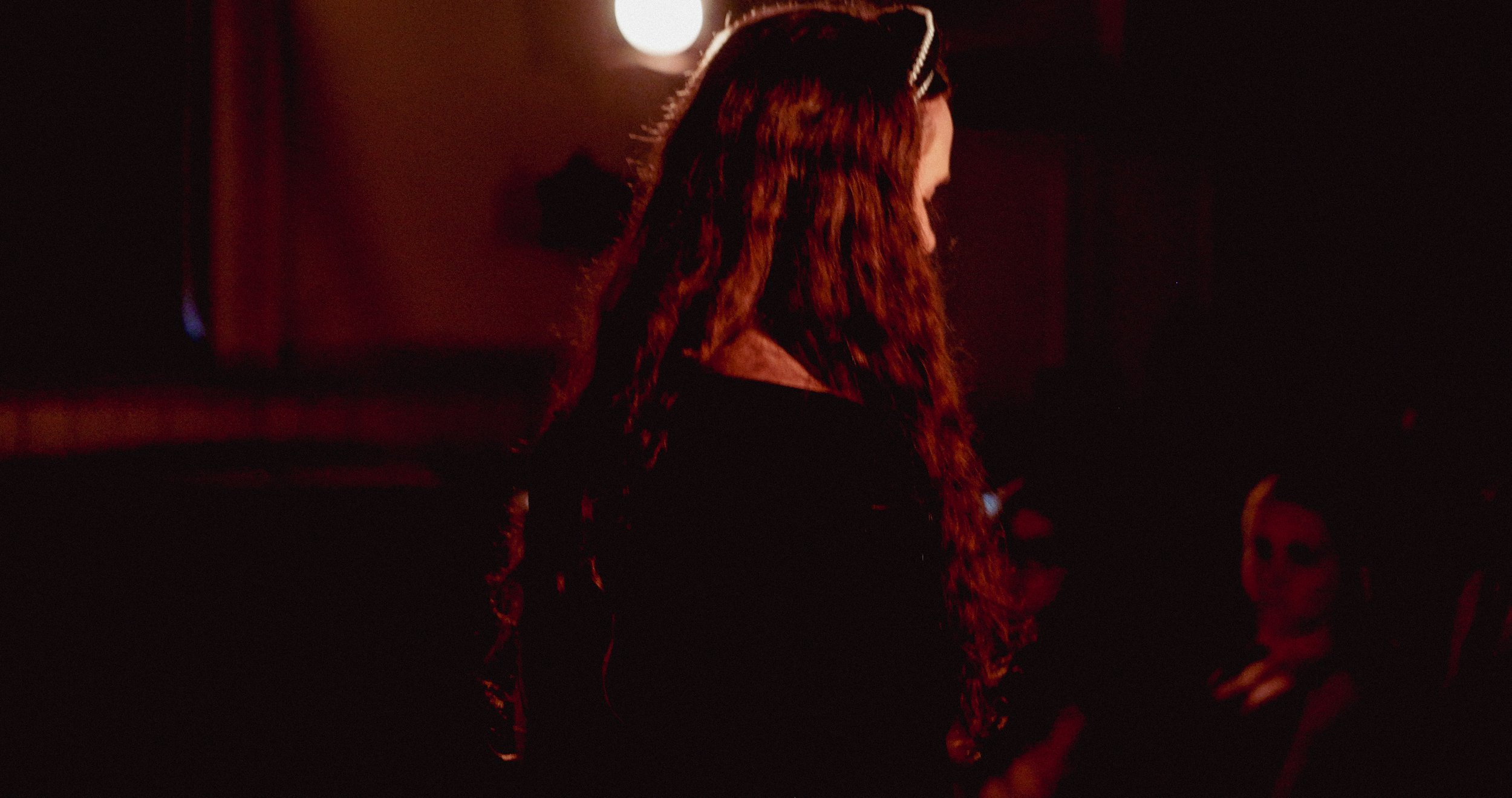 A woman with long, wavy red hair standing in a dimly lit room with red lighting, facing to the right, next to a person wearing glasses and dark clothing.