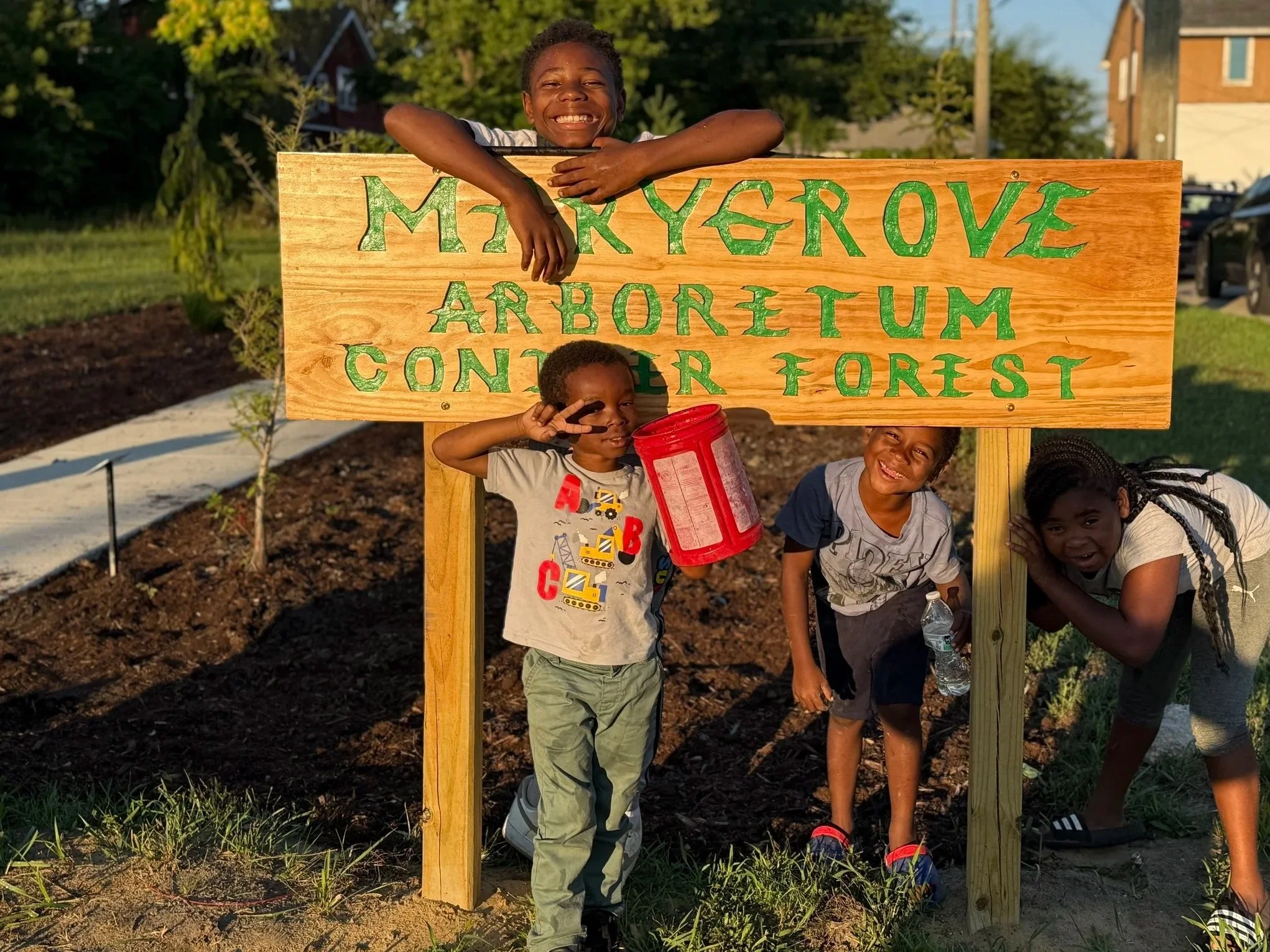 Four children smiling and posing behind a wooden sign that reads 'MakyGrove Arboretum Center for Forest' in a grassy outdoor setting with trees, houses, and parked cars in the background.