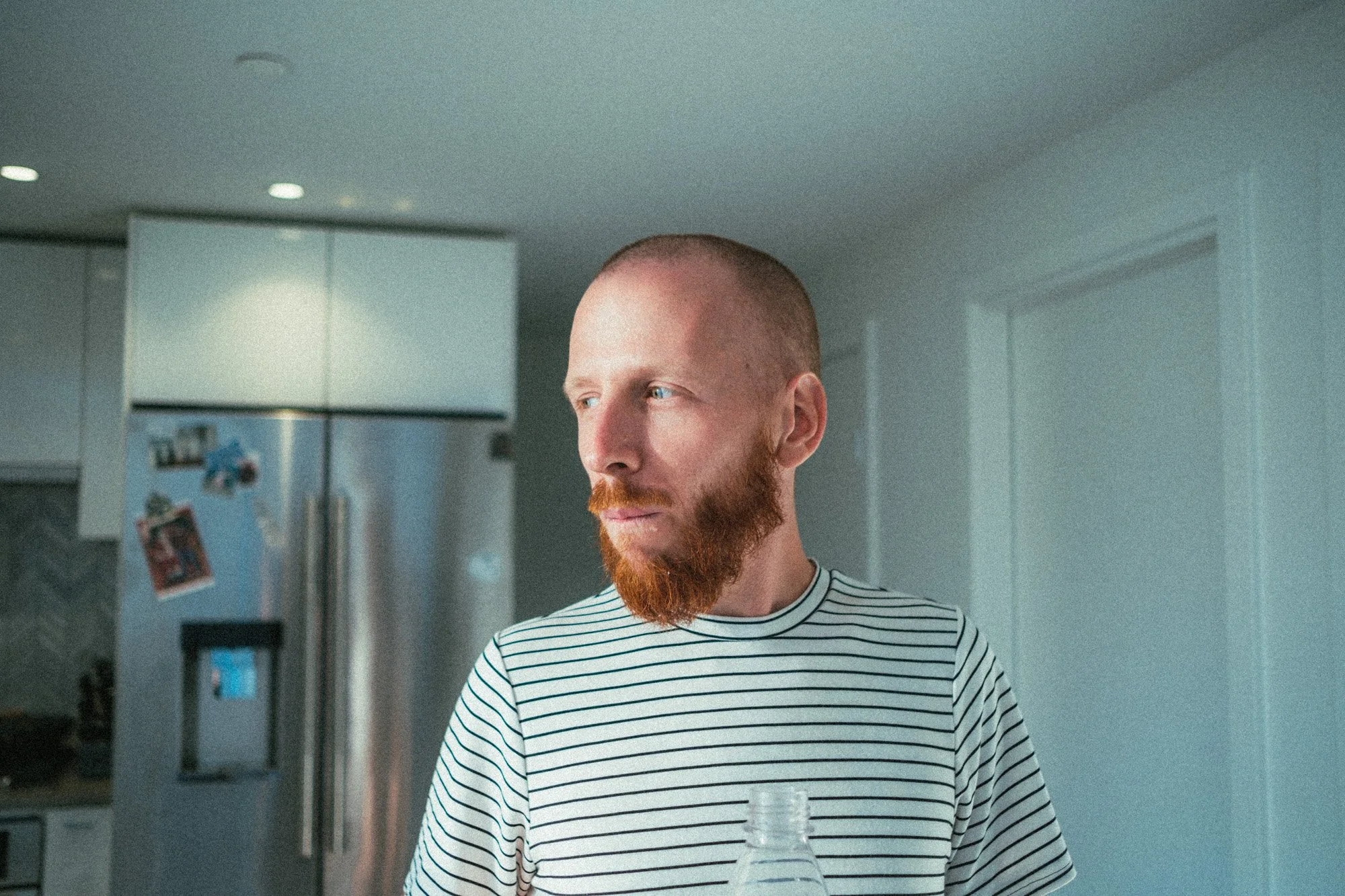 Jeune homme aux cheveux ras et barbe rousse dans une cuisine moderne, portant un t-shirt à rayures et tenant une bouteille en plastique transparente.