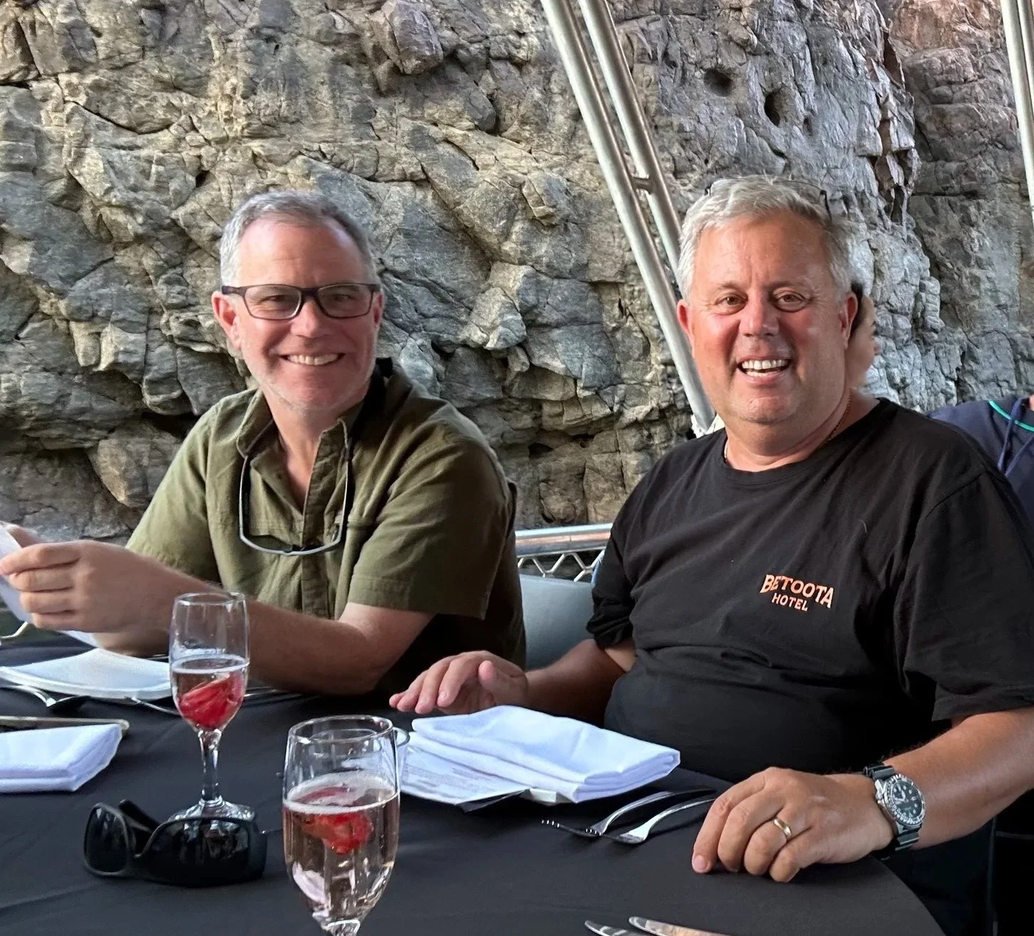 Two smiling men sitting at a table in a restaurant or event space with a rocky wall background, glasses with pink drinks, and white napkins on the table.