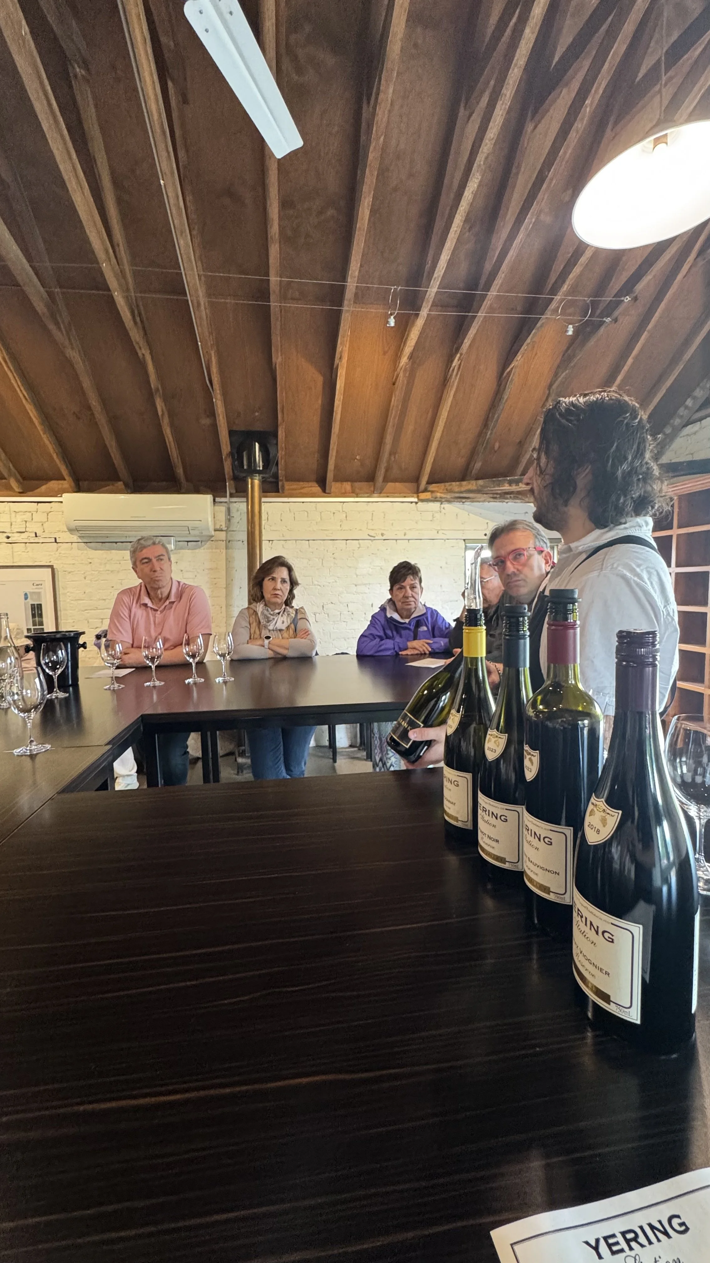 A man holding several bottles of wine is speaking to a group of four women seated at a table in a winery tasting room. The room has a wooden ceiling with a white brick wall in the background.