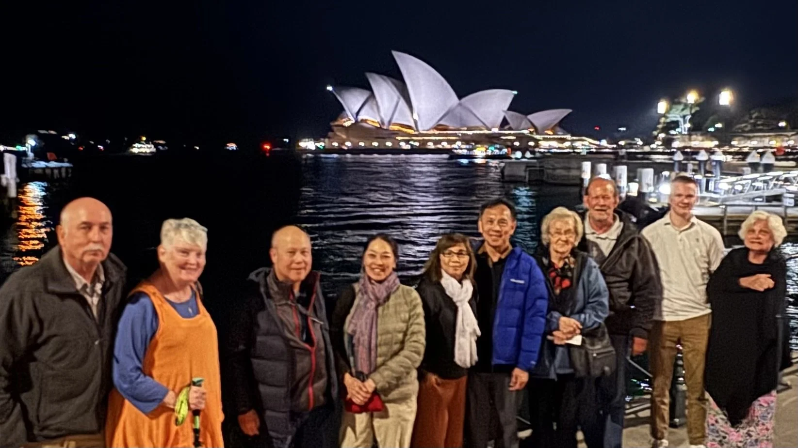 Group of nine people posing for a photo at night with the Sydney Opera House and harbor in the background.