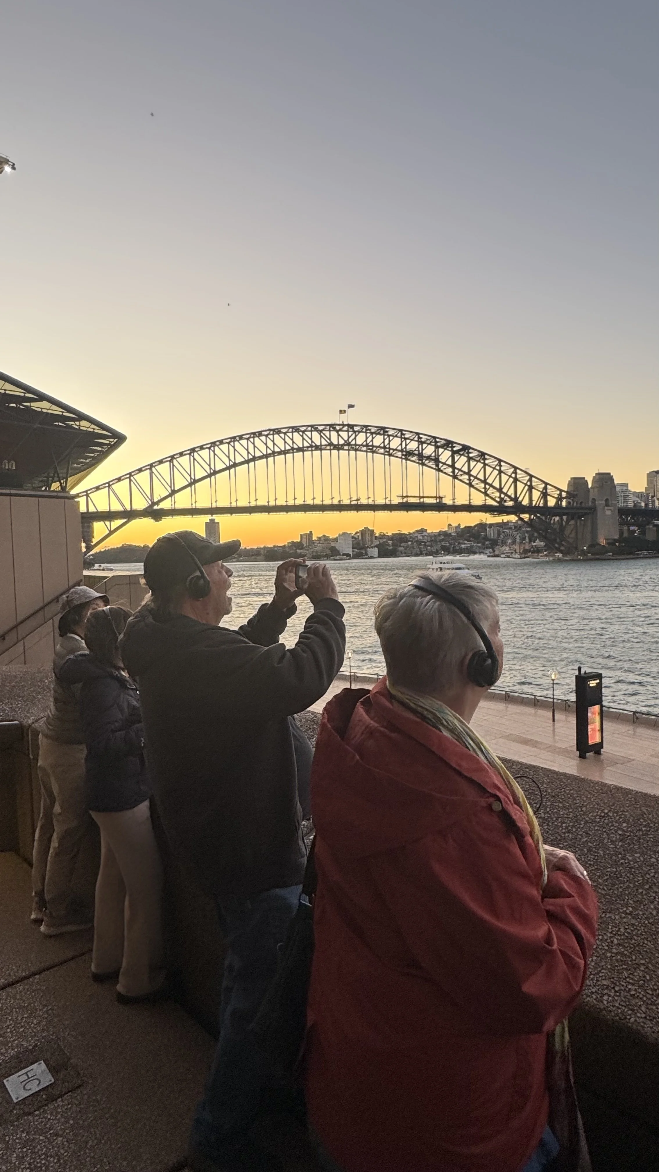 Four people wearing headphones are viewing the Sydney Harbour Bridge at sunset, with city buildings in the background.