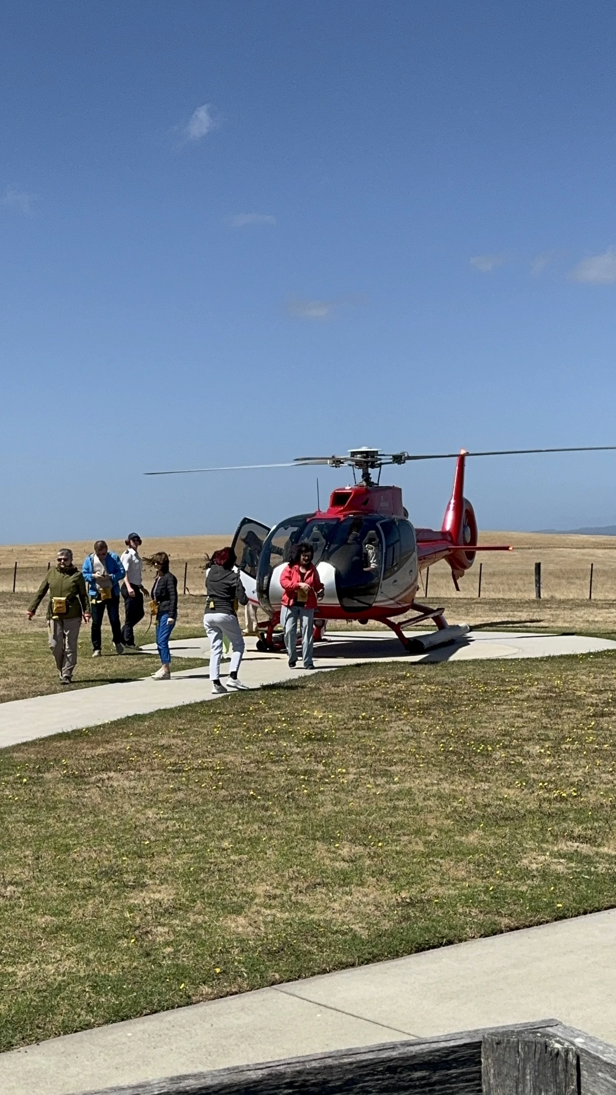 People gather around a red helicopter on a small helipad, with a grassy field and open sky in the background.