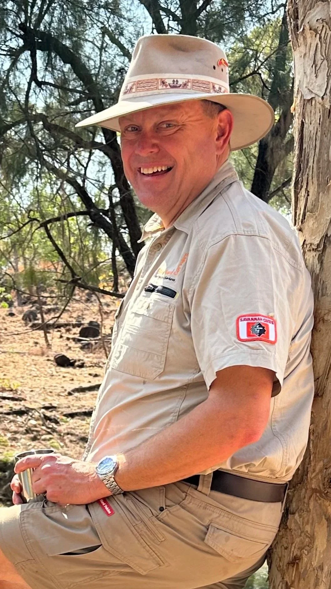 A smiling man in a beige uniform and wide-brimmed hat, sitting outdoors against a tree, with a badge on his shirt and a patch on his sleeve.