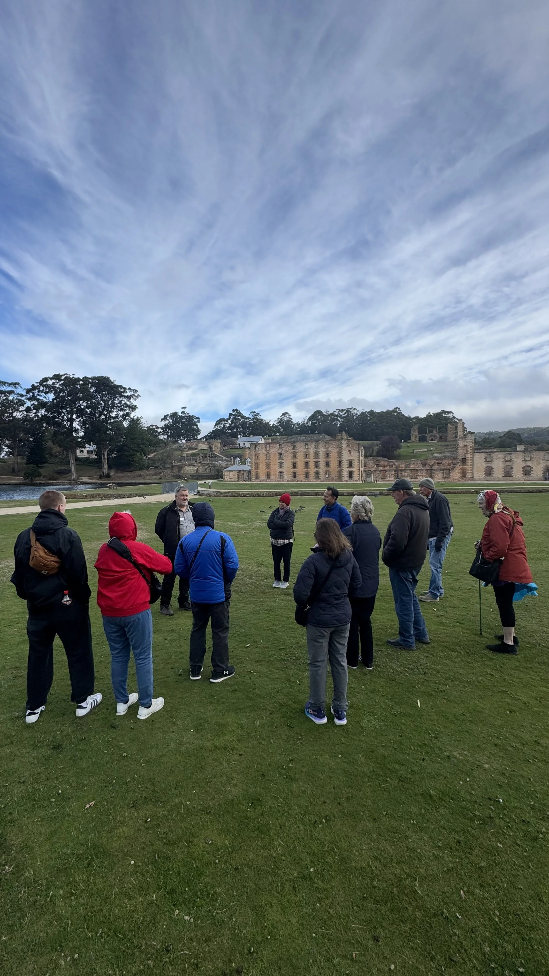 A group of people gathered outdoors on a grassy area, listening to a person speaking. In the background, there are historic ruins and a building, with trees and a cloudy sky overhead.