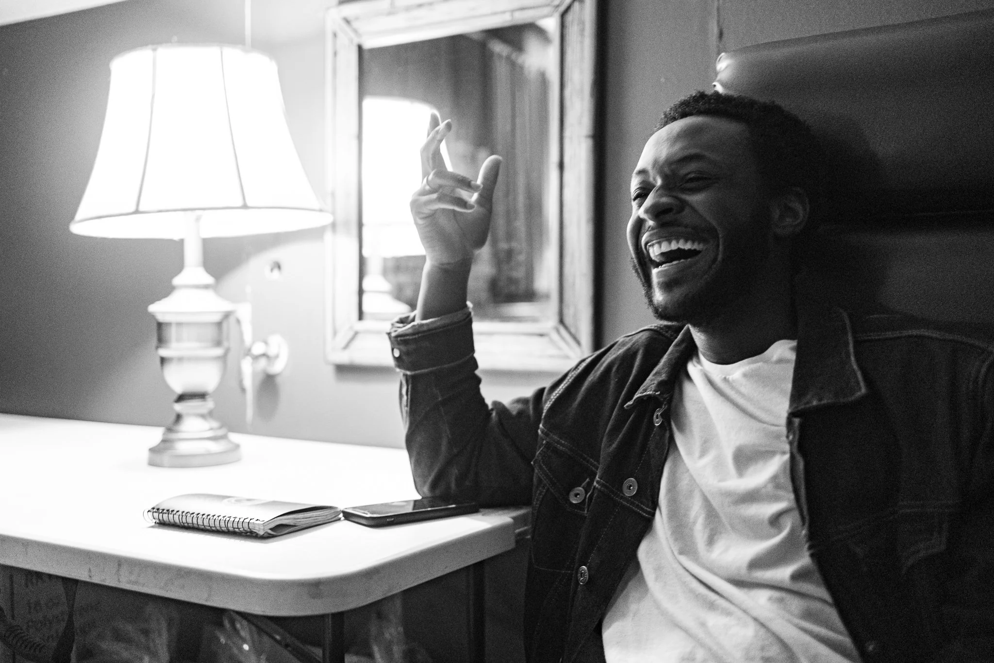 A man sitting at a desk, smiling broadly with eyes closed, gesturing with his right hand, in a room lit by a table lamp, with a notebook and phone on the desk, and a mirror on the wall.
