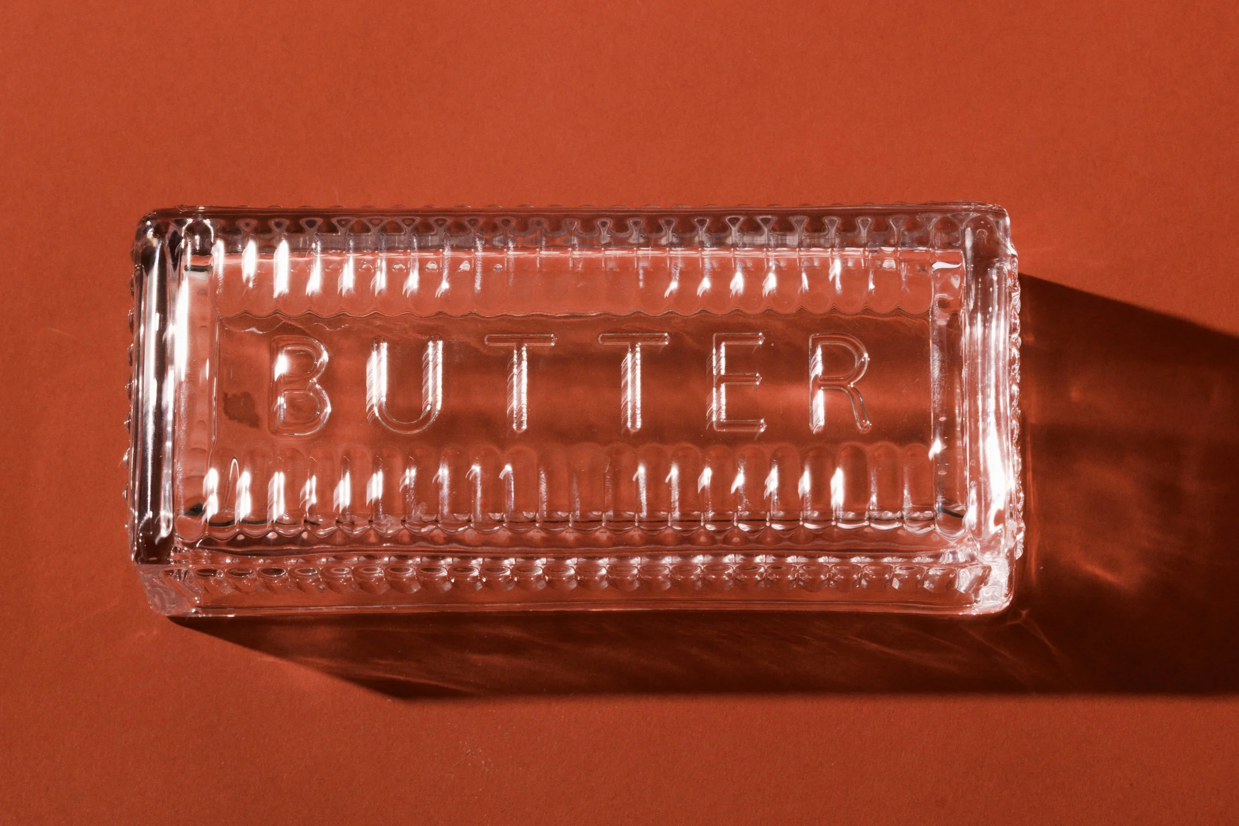 A clear glass block with the word 'BUTTER' engraved on it, placed on a reddish-brown surface with an orange background.