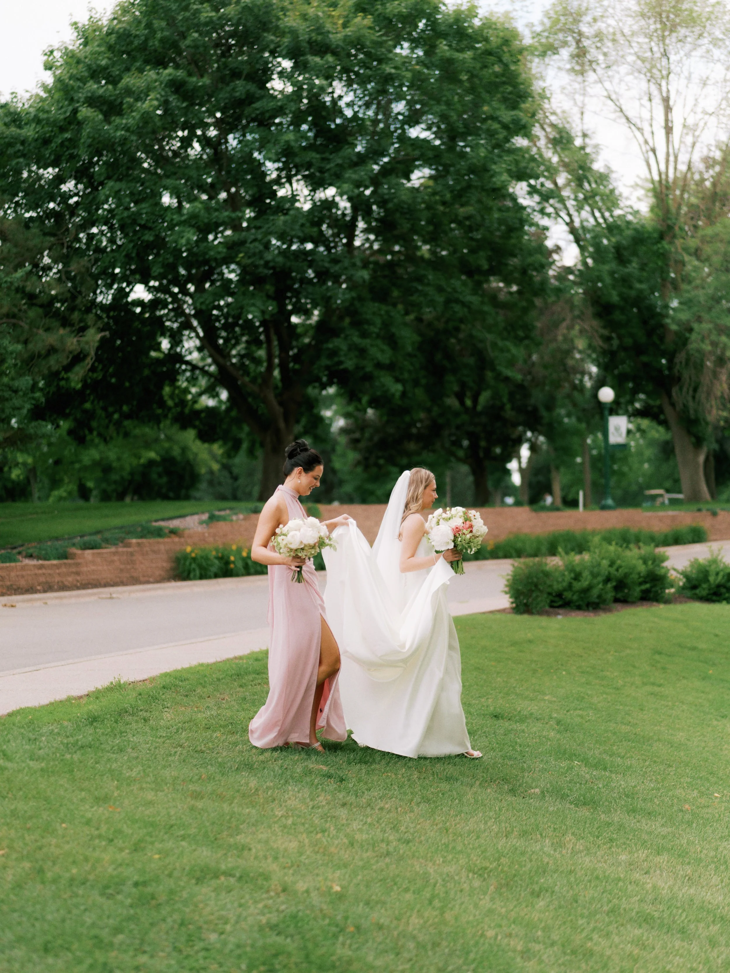 A bride in a white wedding dress holding a bouquet walking on grass, accompanied by bridesmaids in pink dresses holding bouquets, in a park with trees and a walking path.
