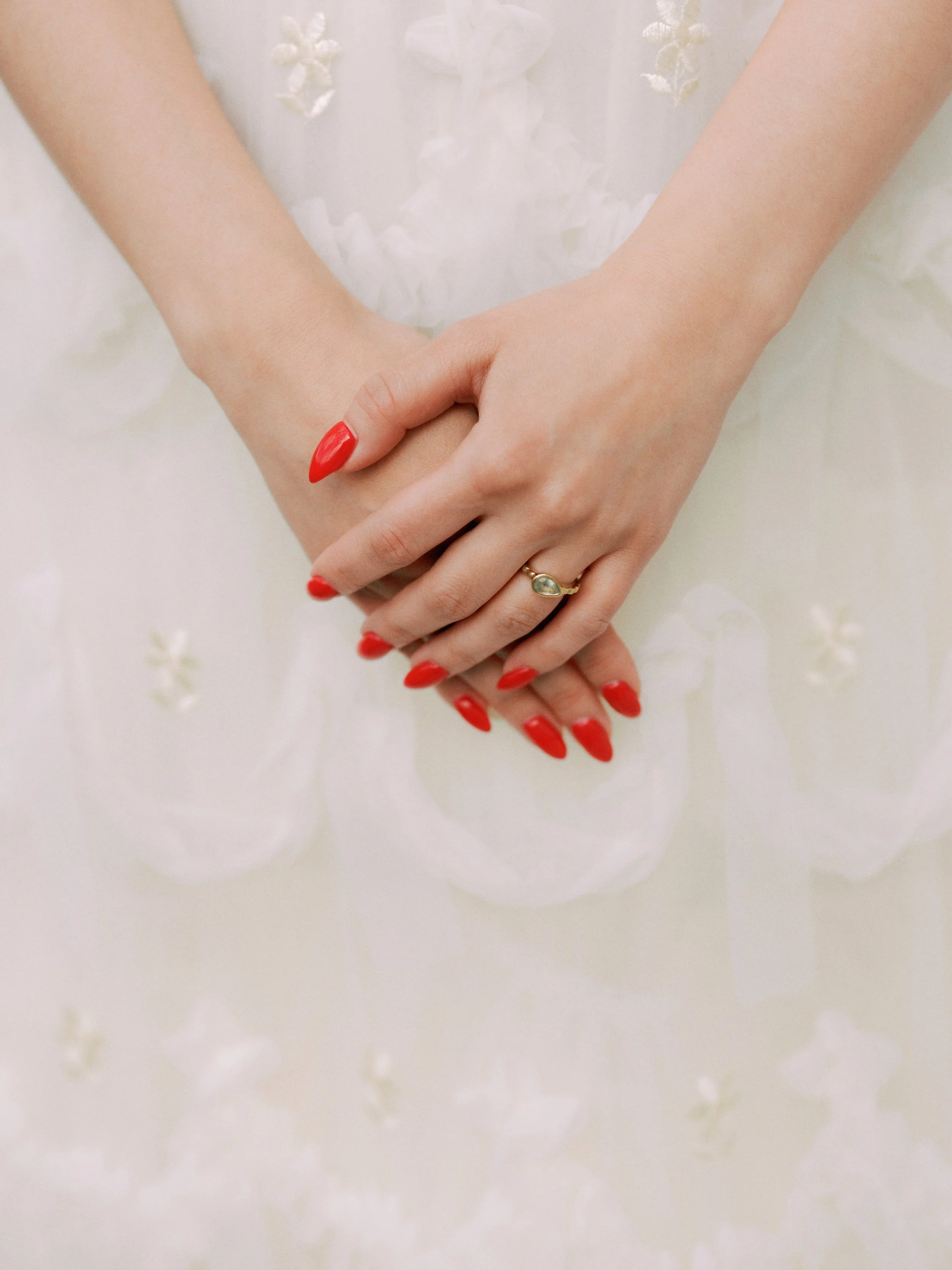 Close-up of a woman's hands with red nail polish, wearing a gold ring with an oval gemstone, resting on a white dress with floral embroidery.