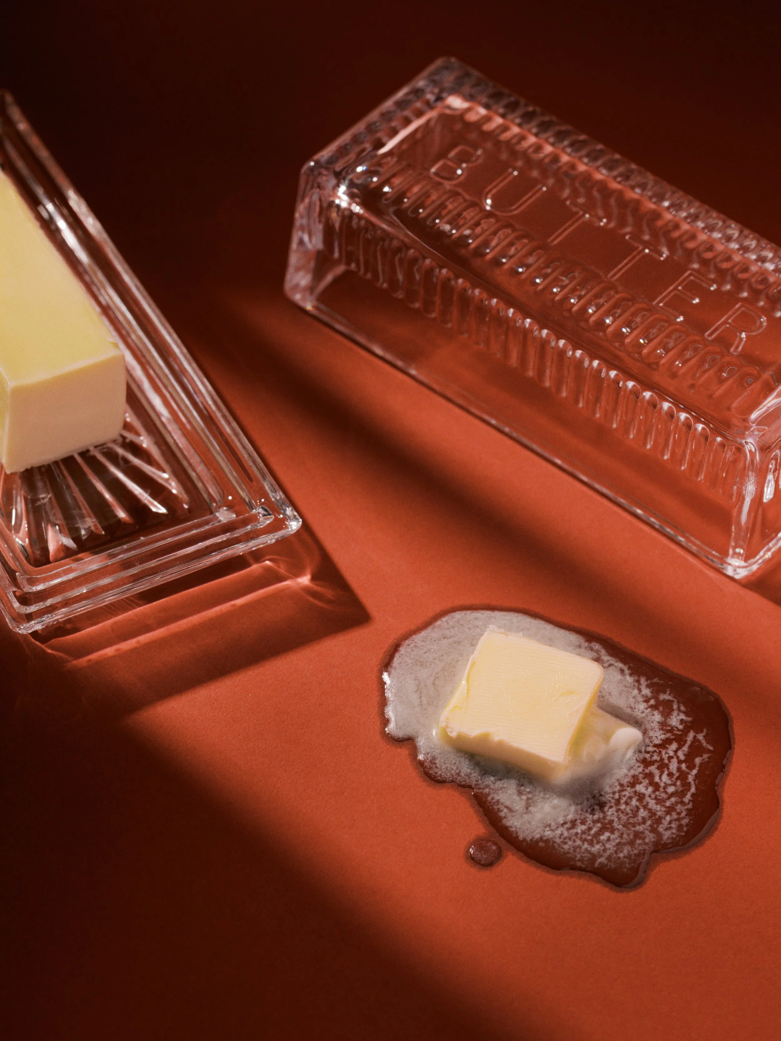 A small glass dish with a pat of butter on top of a dark sauce, next to a clear plastic box of butter on a copper-colored surface.