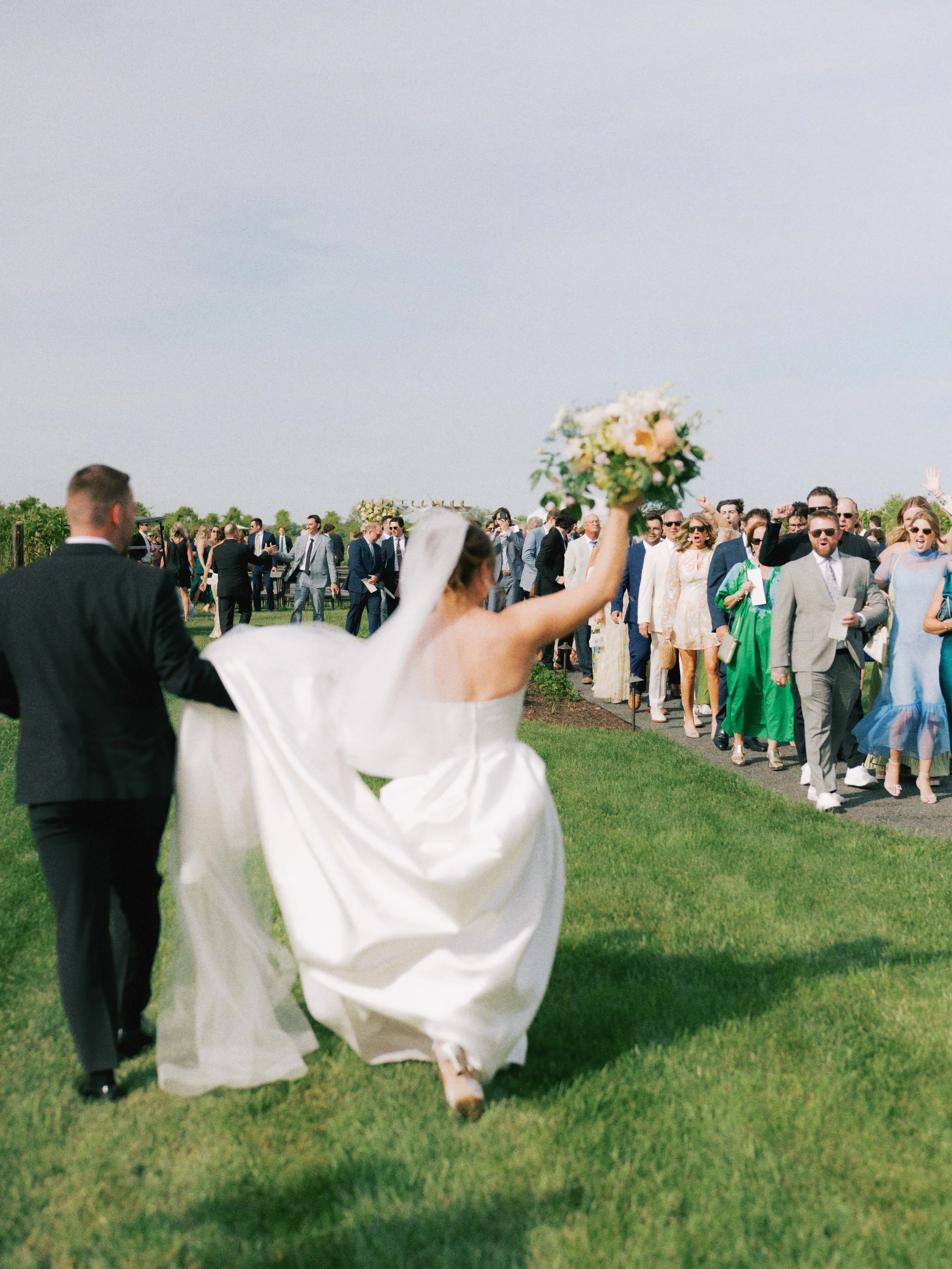Bride holding a bouquet walking on grass at outdoor wedding with guests in the background.