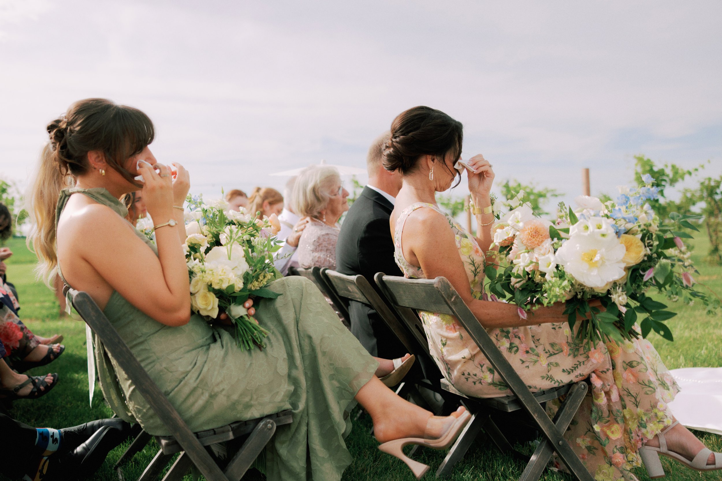 A group of people sitting outdoors on folding chairs at a wedding, holding facial tissue to their faces during a tearful moment, surrounded by floral arrangements and greenery.