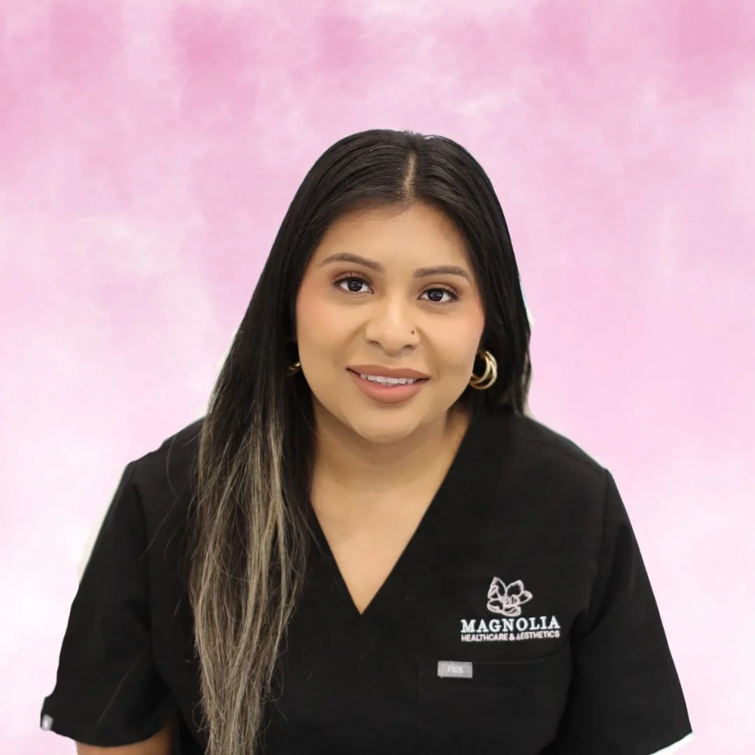 A woman with long, wavy hair smiling, wearing a black medical uniform with 'Magnolia Healthcare & Aesthetics' logo, arms crossed, standing against a pink background.