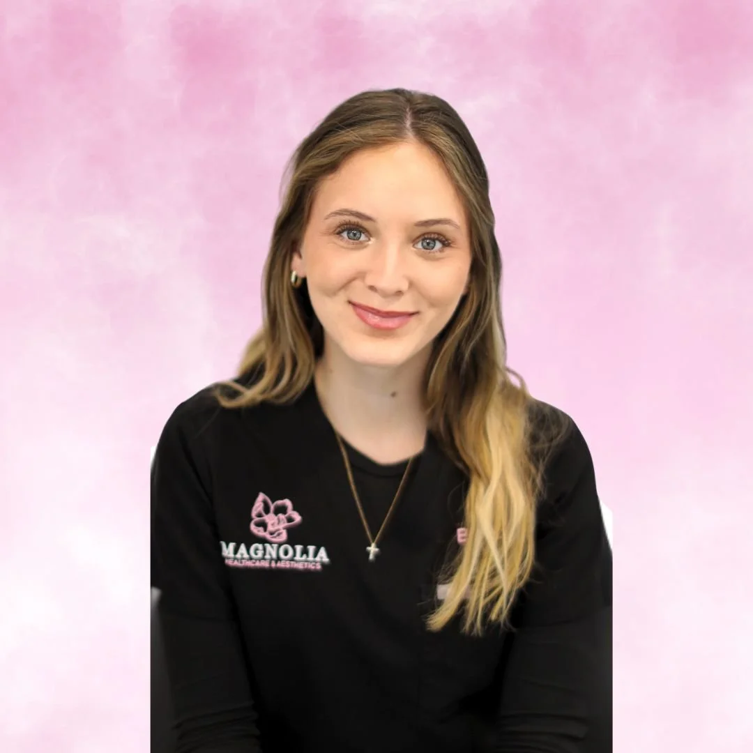 A woman with blonde hair and blue eyes smiling, wearing a black jacket with pink and white embroidered text and logos, standing against a pink background.