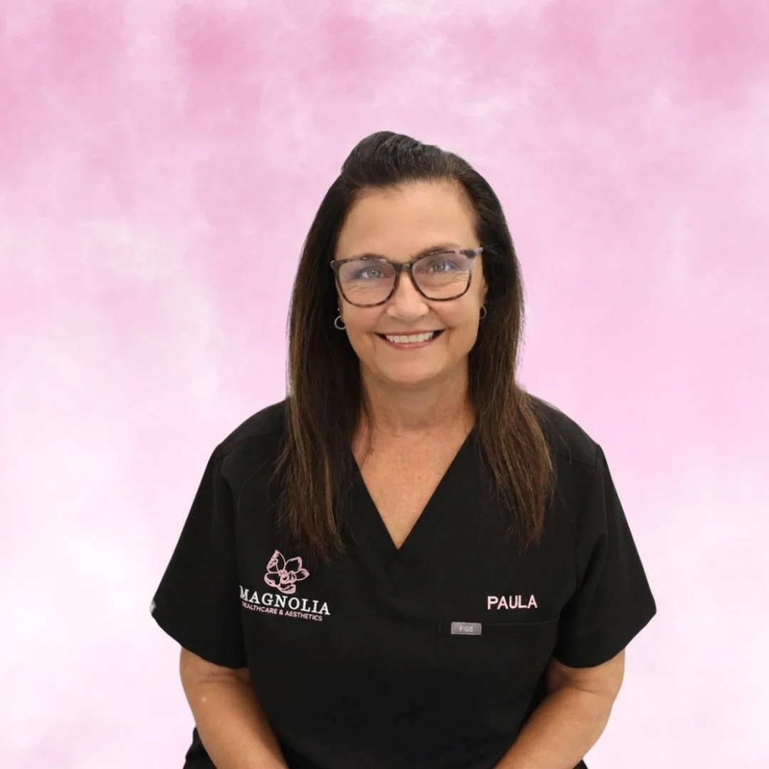 A woman with shoulder-length brown hair, glasses, and a black uniform with a pink logo that reads 'Magnolia Healthcare & Aesthetics' and a name tag that says 'Paula'. She is smiling and standing against a pink background.