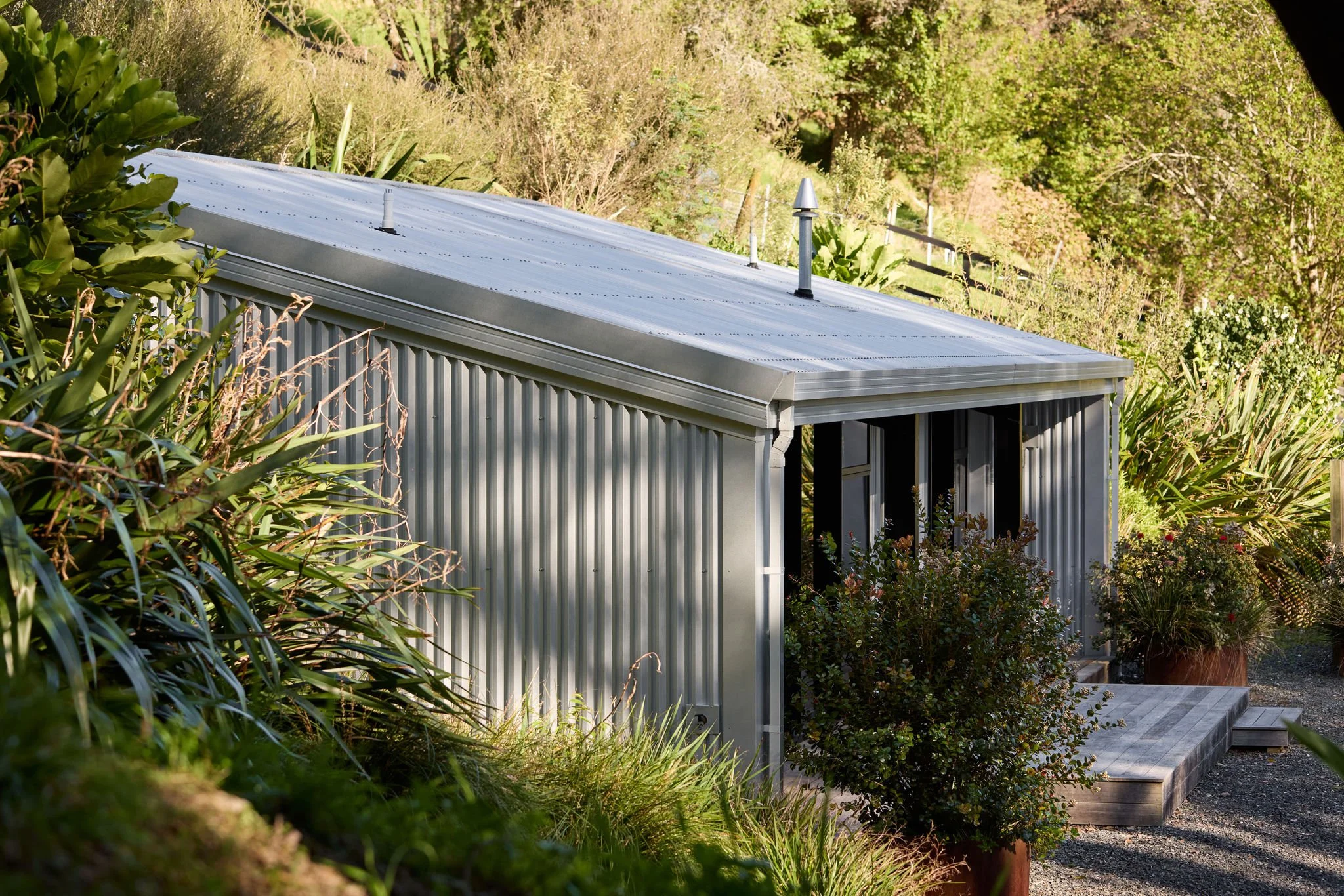 Metal shed with a sloped roof, surrounded by lush green plants and trees, and a wooden platform at the front.