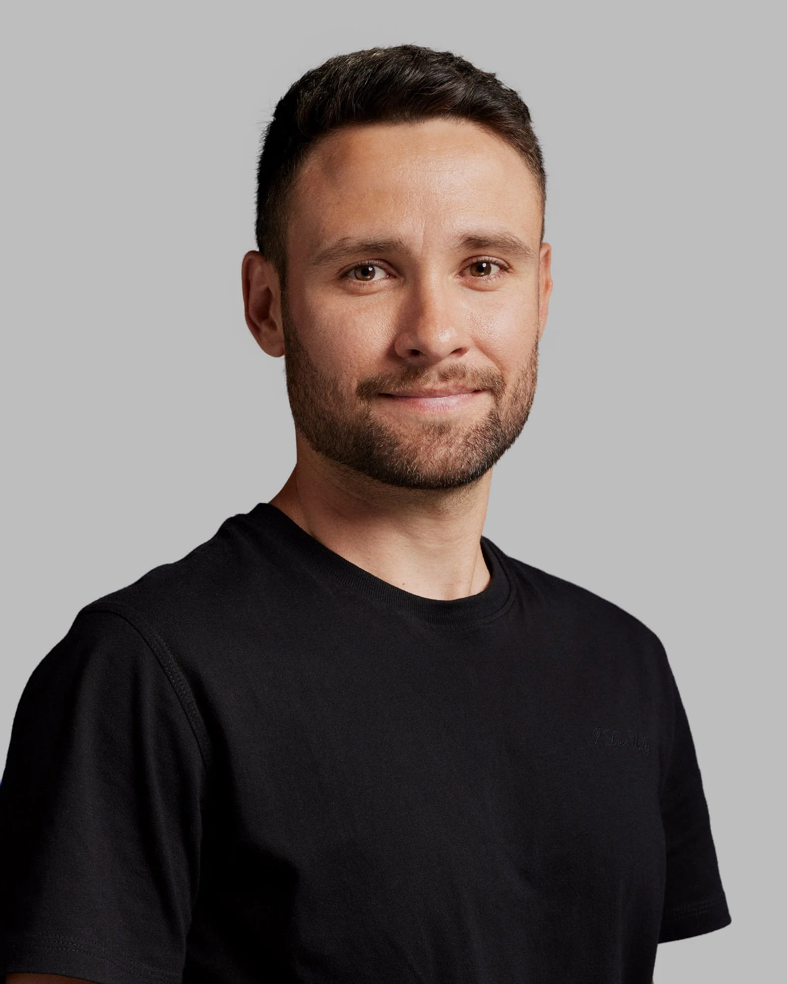 Portrait of a young man with dark hair and a beard, smiling slightly, wearing a black T-shirt, against a gray background.