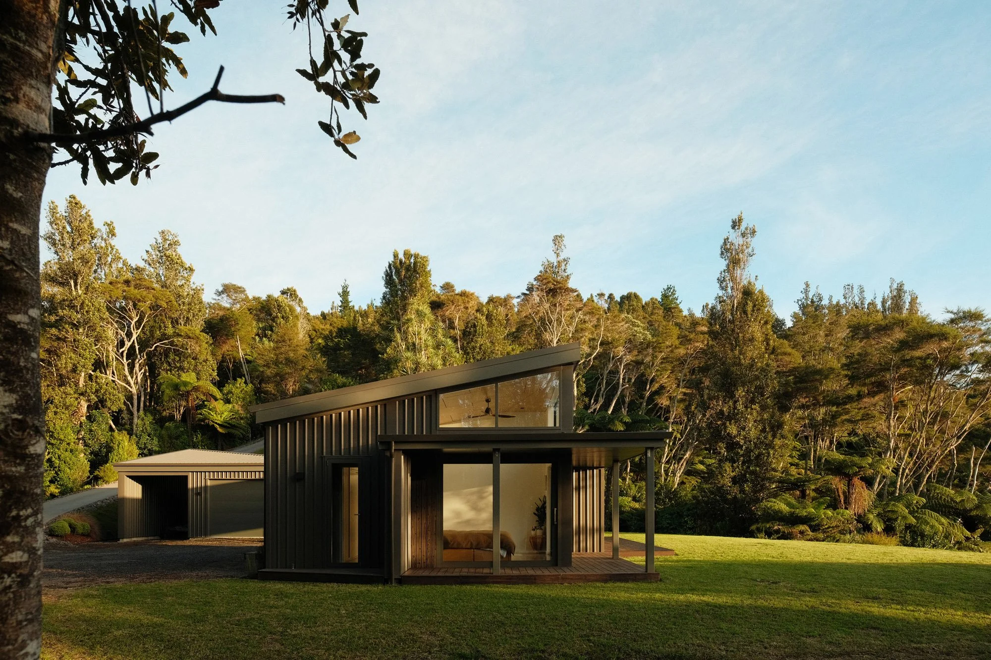 Modern house with large glass windows and vertical metal siding, situated on a lush green lawn, surrounded by trees and a hillside in the background under a partly cloudy sky.