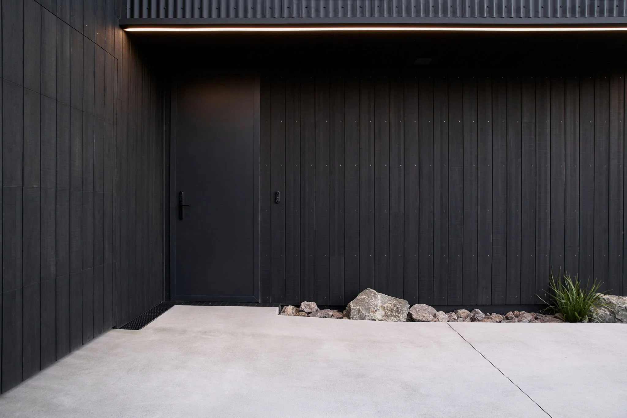 Modern black building exterior with wooden paneling, a closed door, and minimal landscaping with rocks and a small plant.