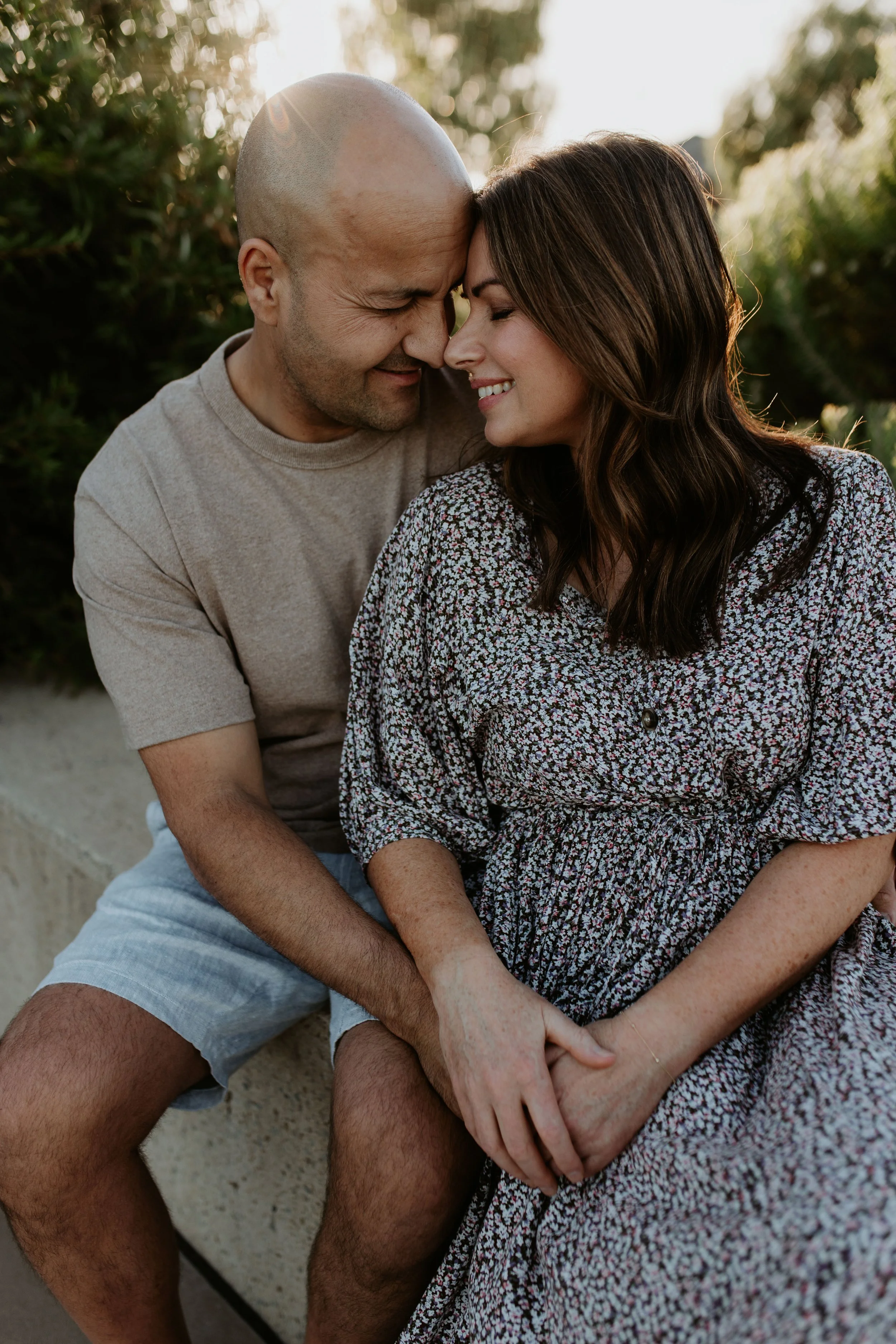 A couple sitting close together outdoors with foreheads touching, smiling, during sunset.