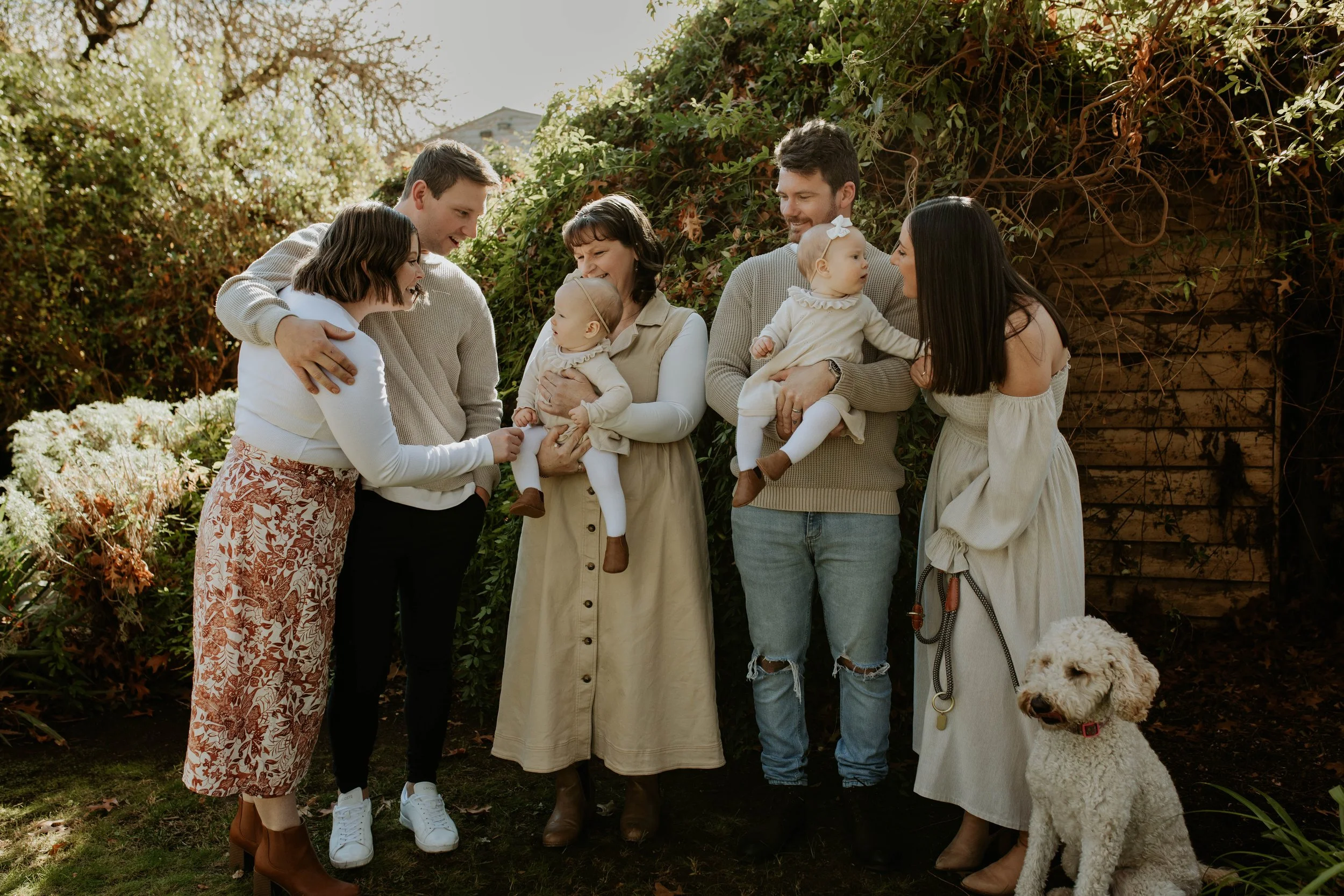 Family gathered outdoors at a garden, smiling and talking, with two young children, a dog, and a background of foliage and a wooden wall.