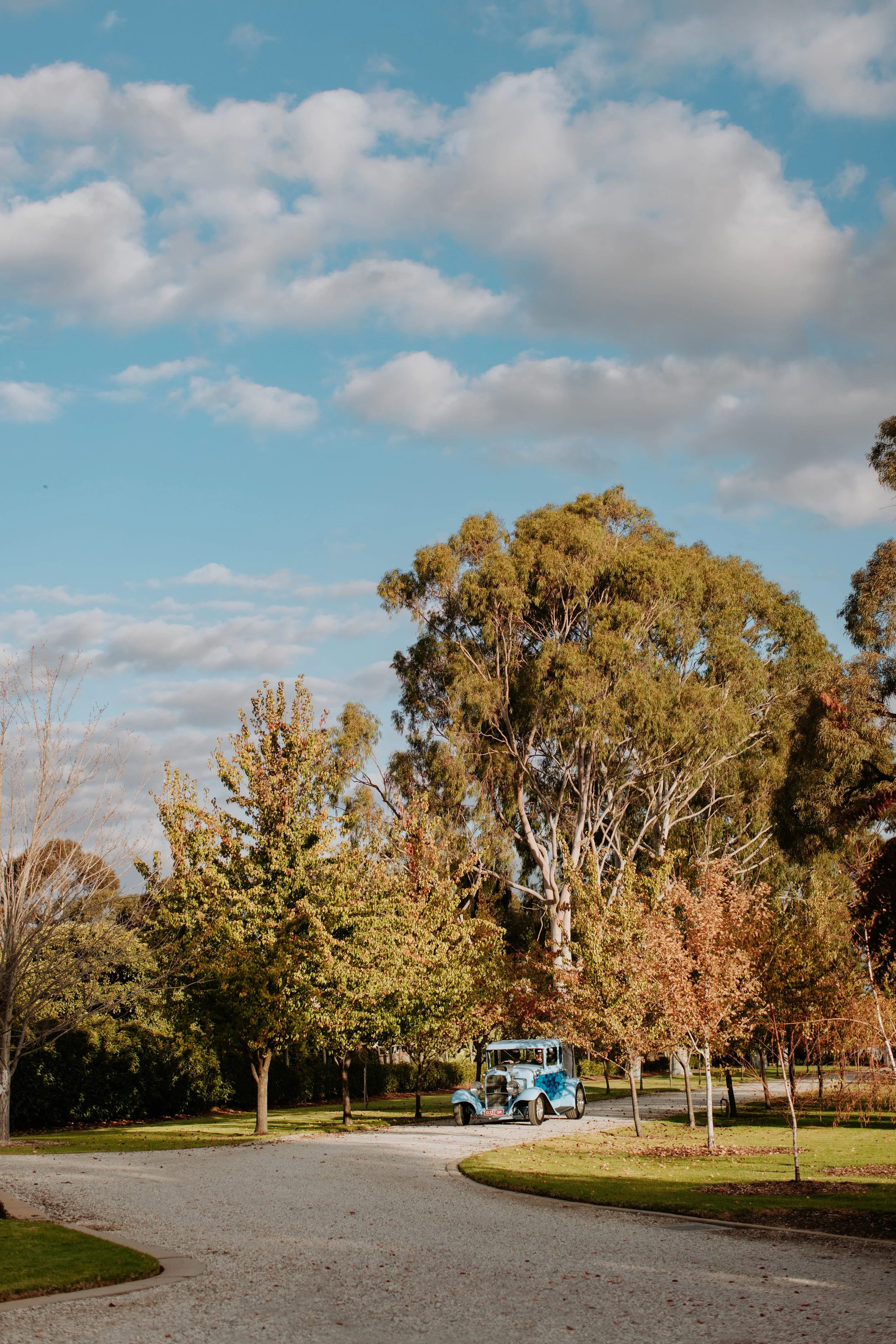 A vintage blue car parked on a curved gravel driveway surrounded by trees with autumn foliage under a cloudy sky.