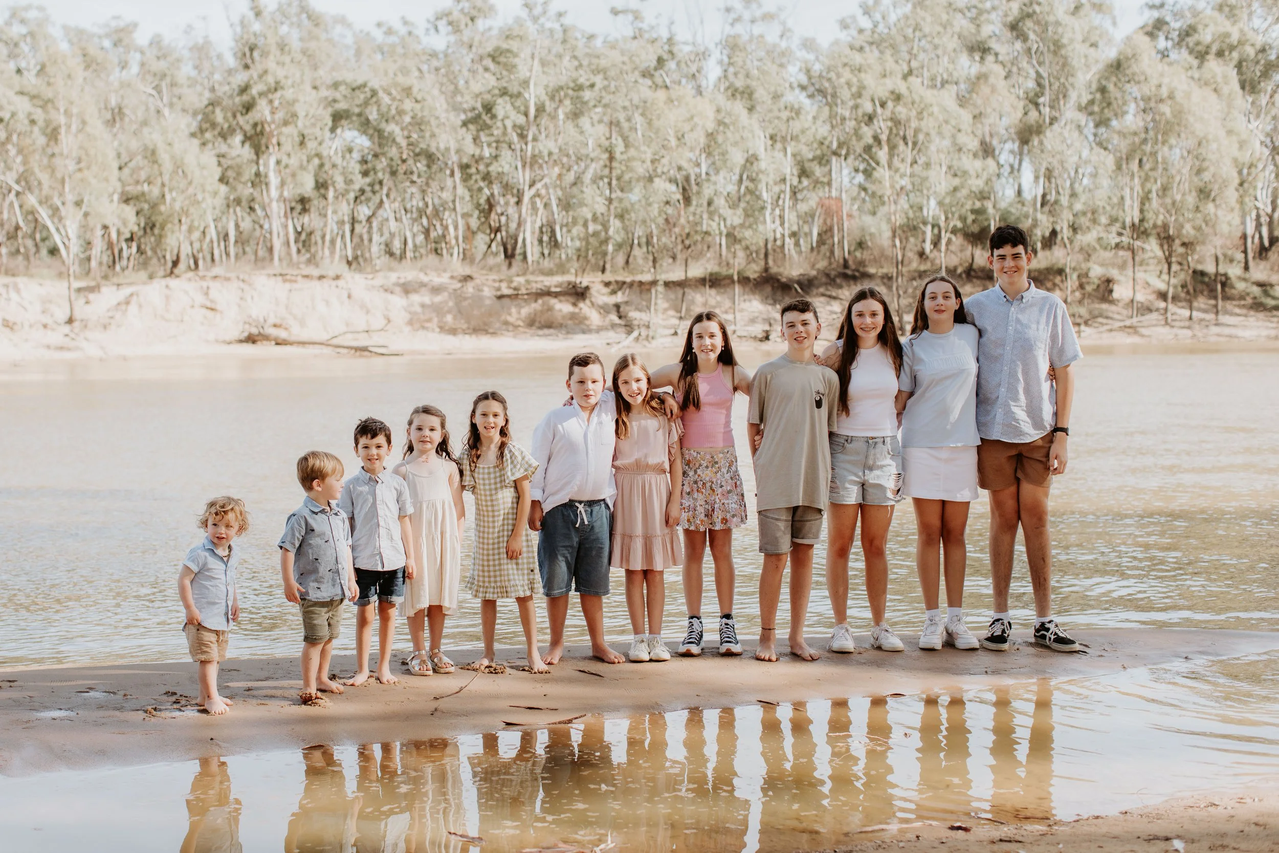 Group of children and teenagers standing on a sandy beach near a body of water, with trees and a hill in the background.