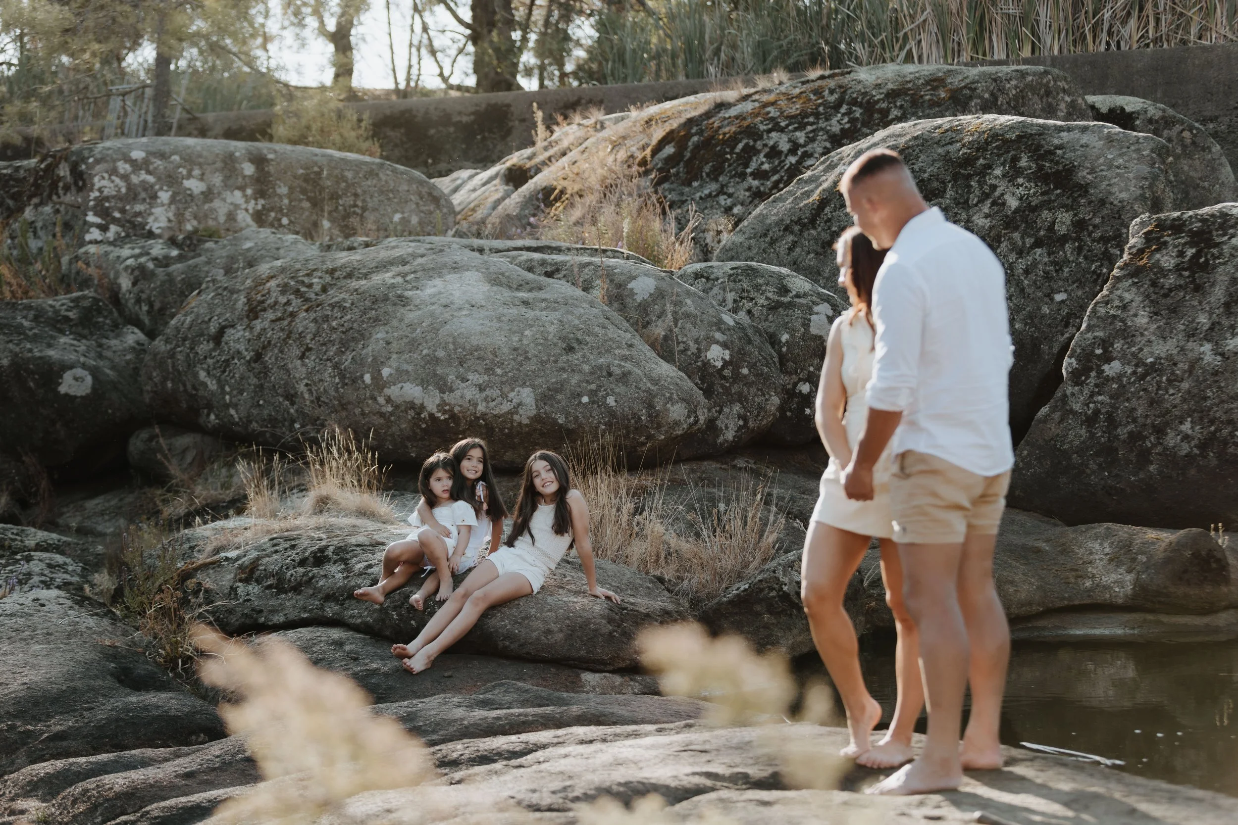 A family of five, including two young children and two adults, standing and sitting on rocks near a small body of water in a natural setting with large boulders and dry grass.
