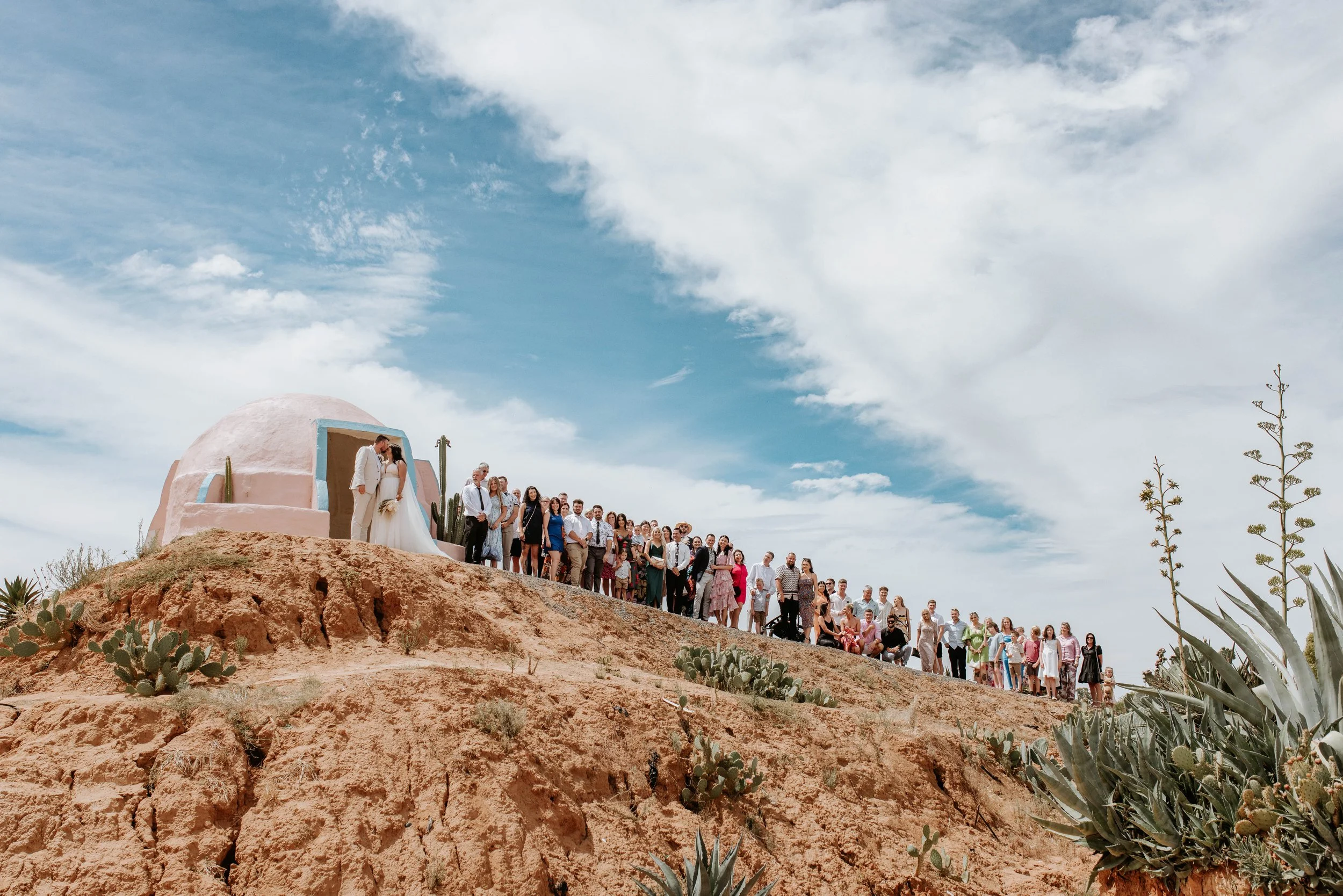 A wedding ceremony with a bride and groom kissing under an adobe-style building on a hilltop, surrounded by wedding guests, desert plants, and cacti, with a partly cloudy sky in the background.