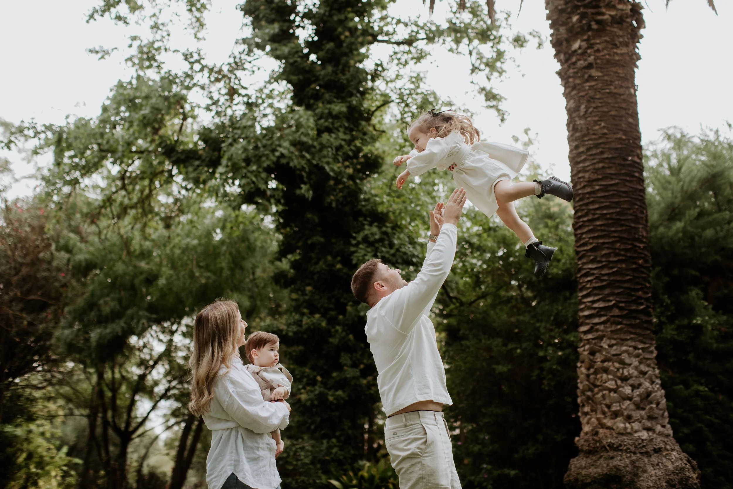 A man is throwing a young girl in the air while a woman holds a baby in her arms, all outdoors among tall trees.