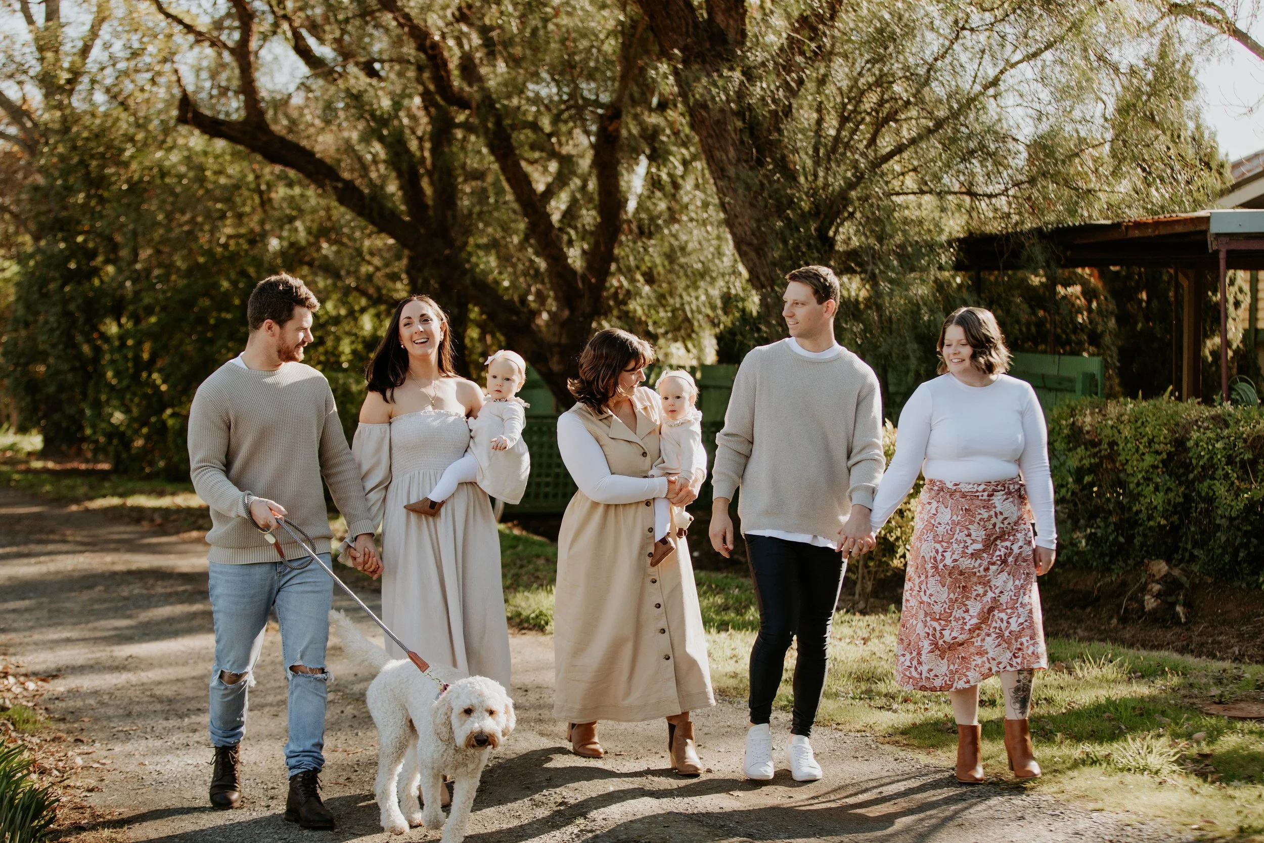 A family walking outdoors in a sunny park, holding hands and smiling, with two young children and a dog.