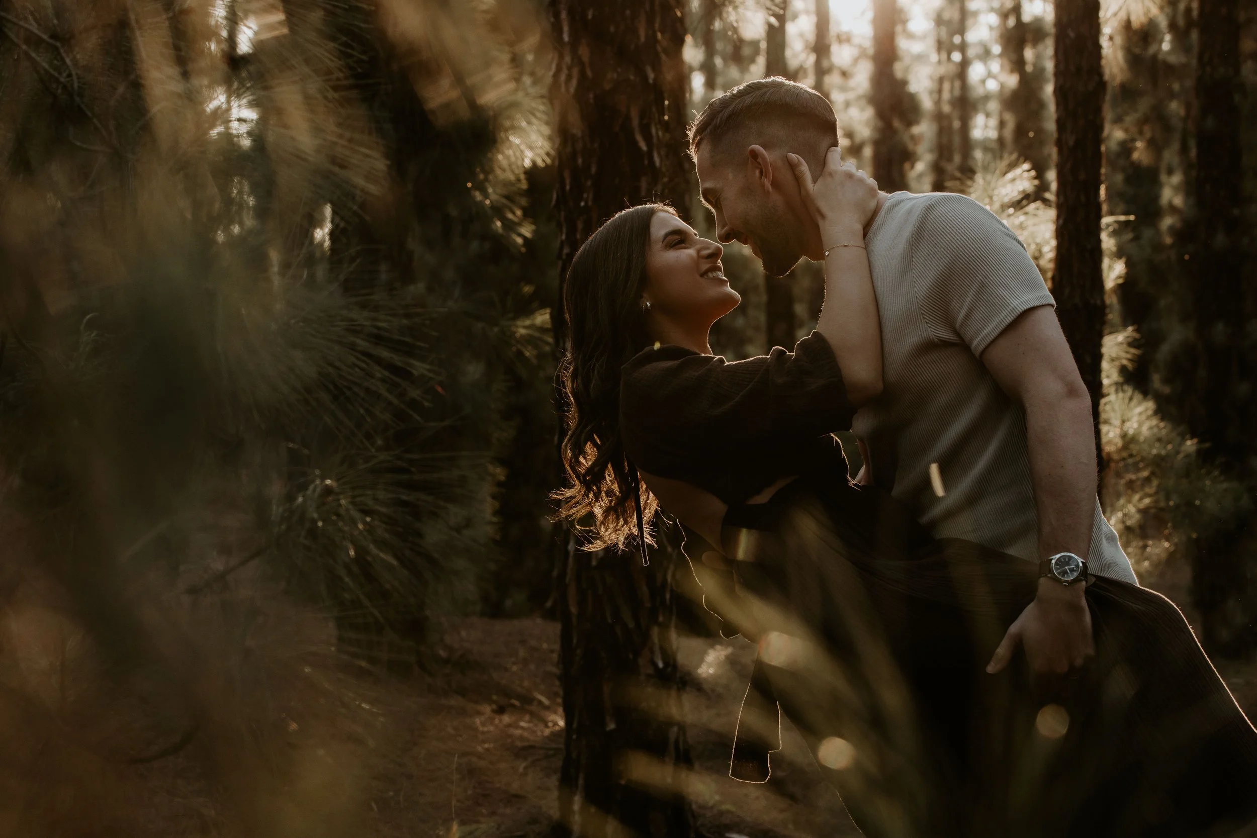 A couple sharing a tender moment in a forest at sunset, with trees and warm light in the background.