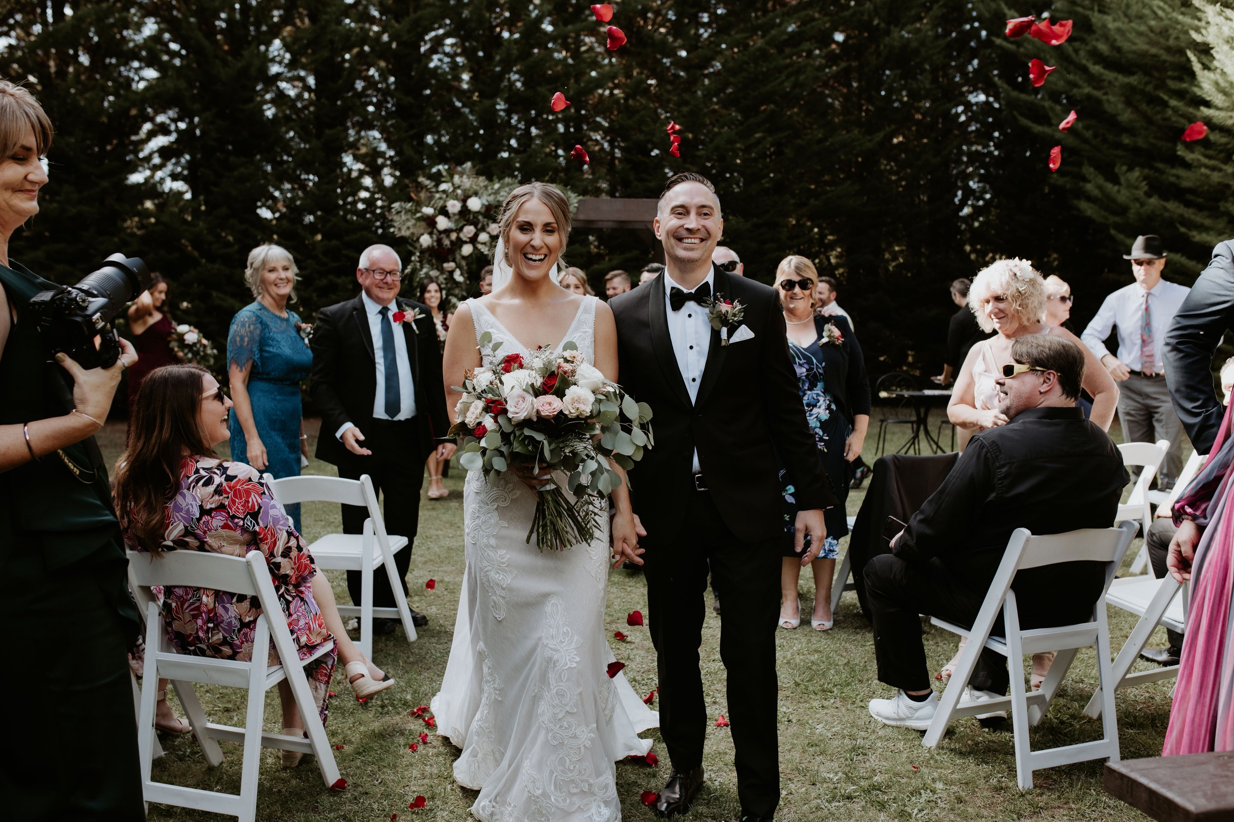 Bride and groom walking down the aisle at their outdoor wedding ceremony surrounded by seated and standing guests, with rose petals falling in the background.
