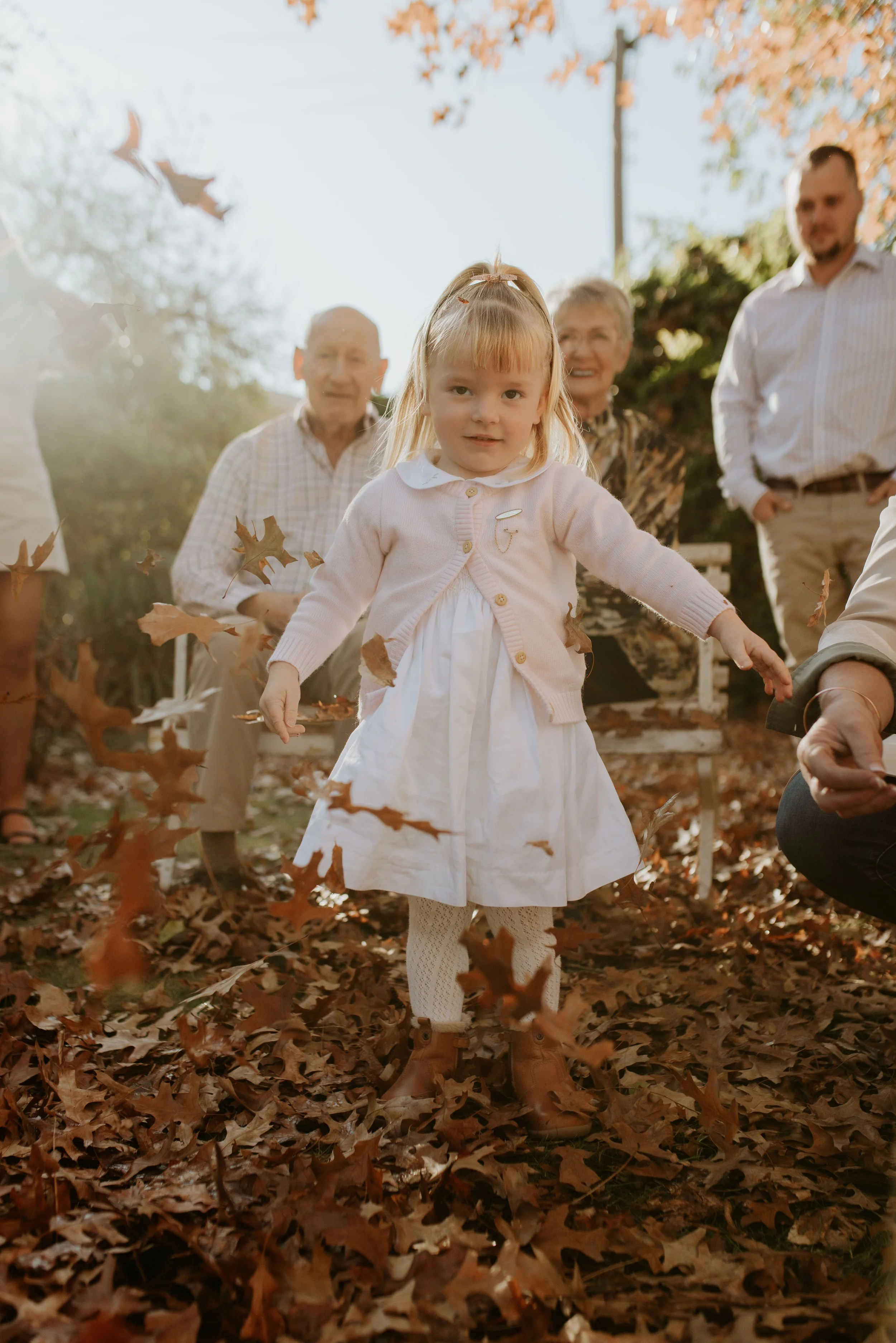 Little girl in a white dress and pink cardigan standing among falling autumn leaves, with family members watching behind her.