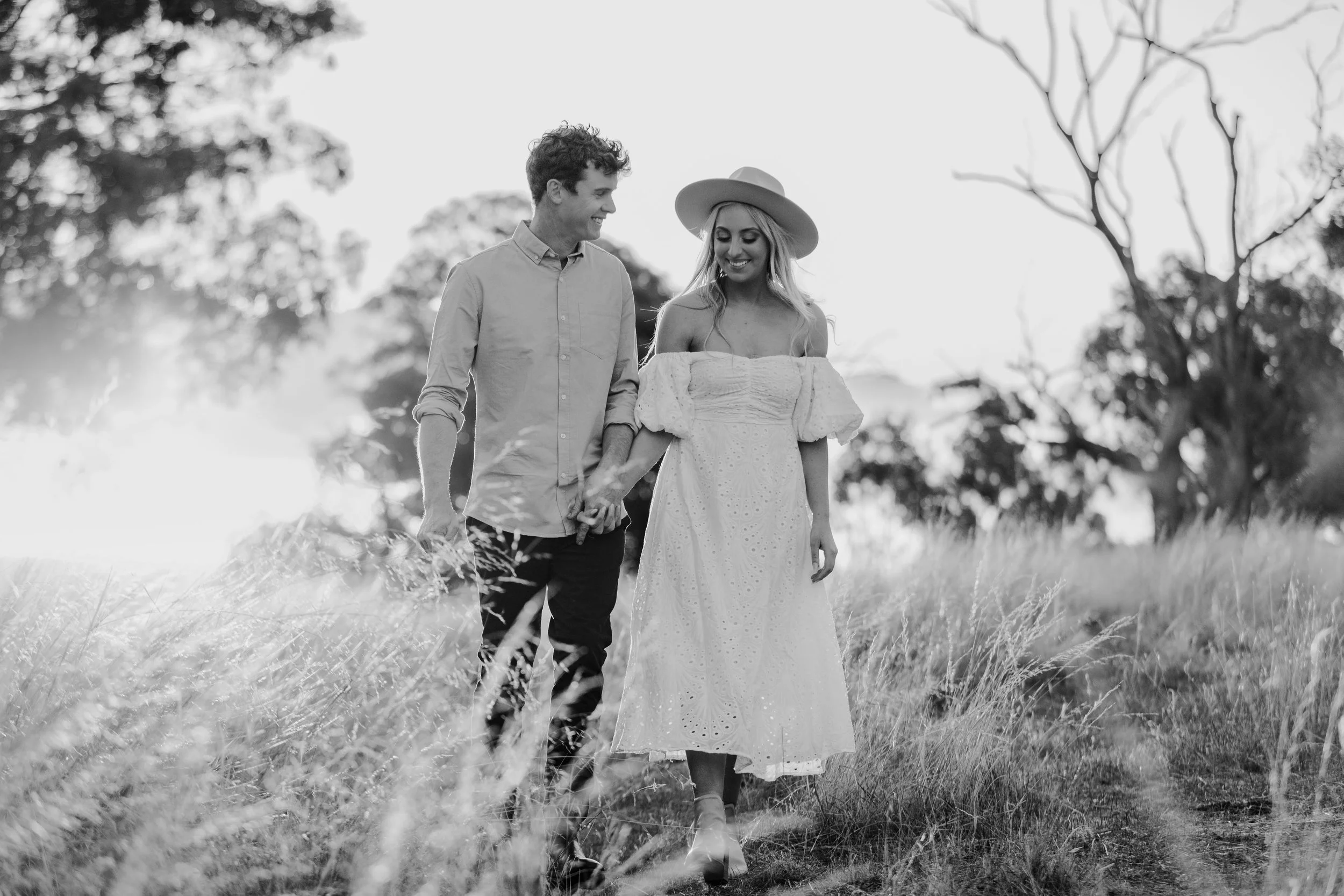 A black and white photo of a couple walking hand in hand through a grassy field, smiling at each other. The woman is wearing a wide-brimmed hat and a long, off-the-shoulder dress. The man is dressed casually in a button-up shirt and pants. Trees are