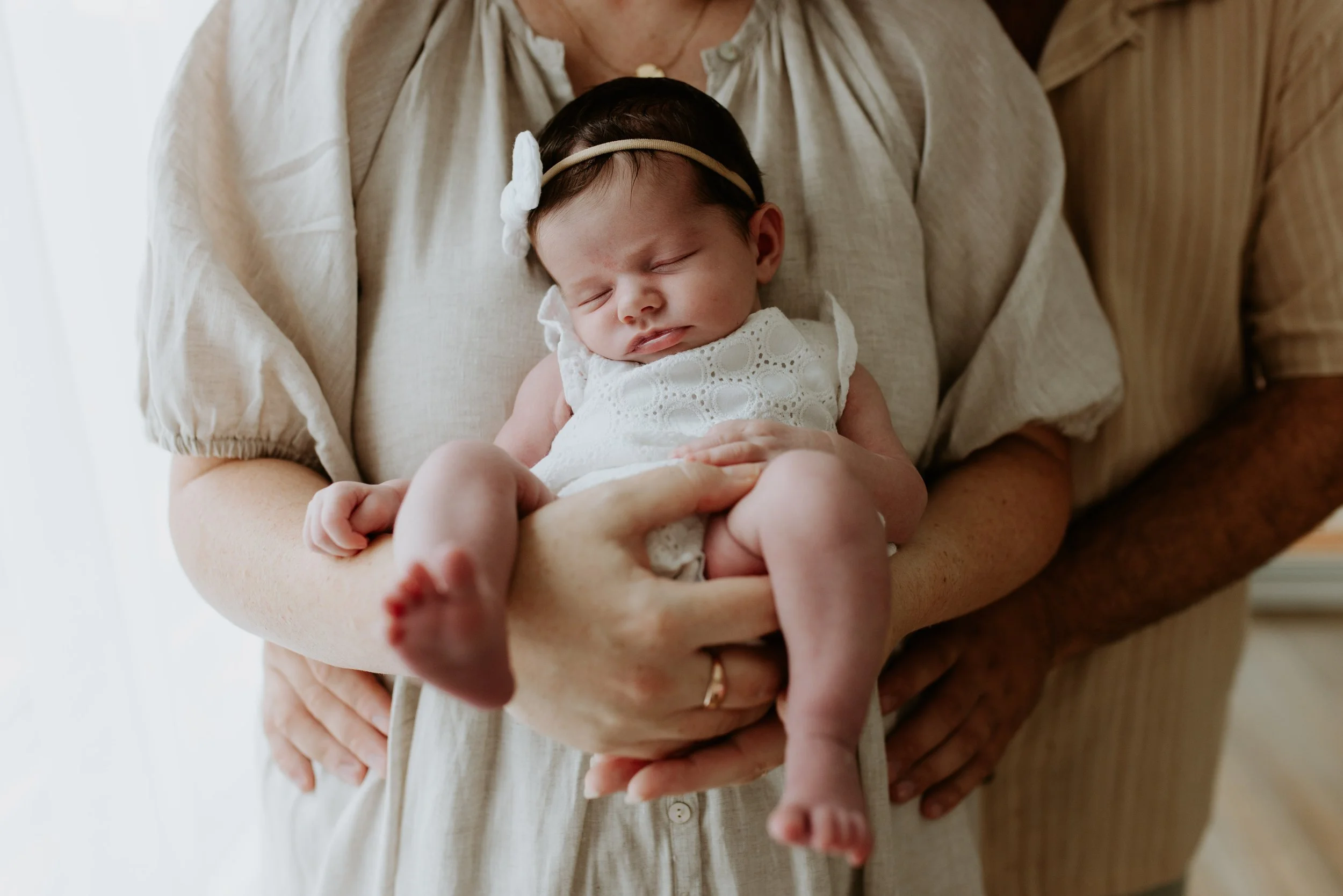 A person gently holding a sleeping baby girl dressed in white, with the baby's head resting on the person's arm and her legs hanging down, with an adult supporting her from behind.