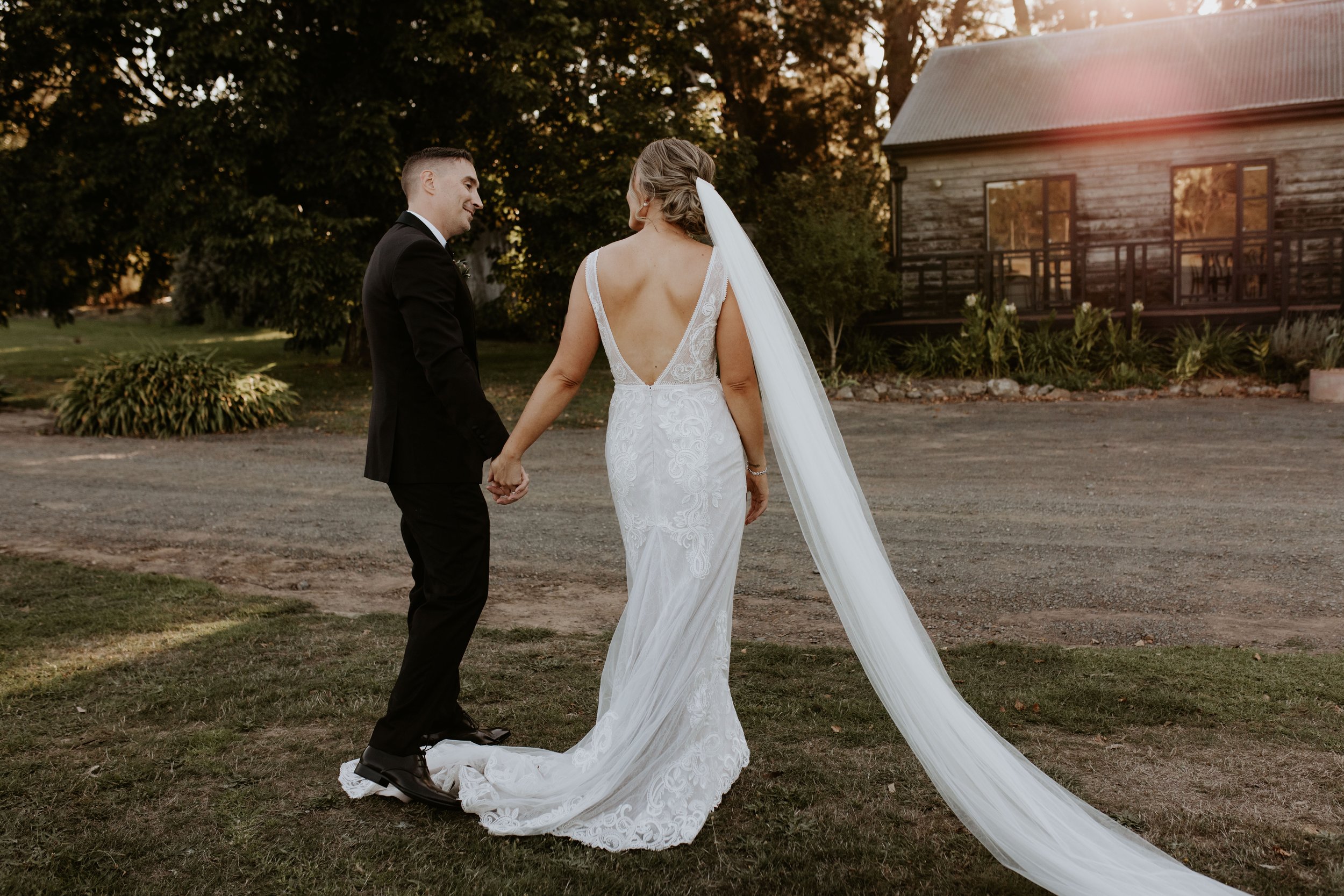 A bride and groom holding hands during their outdoor wedding at sunset, with a rustic barn in the background.