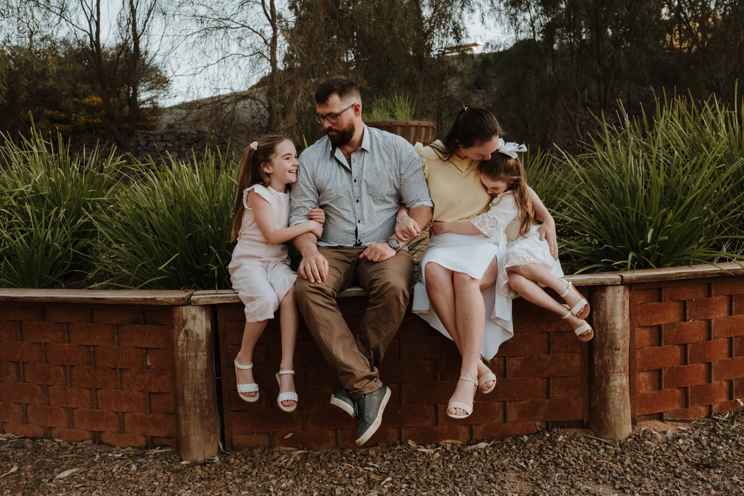 A family of four sitting on a brick and wooden ledge outdoors during autumn, smiling and enjoying each other's company. The family includes a man, a woman, and two young girls.