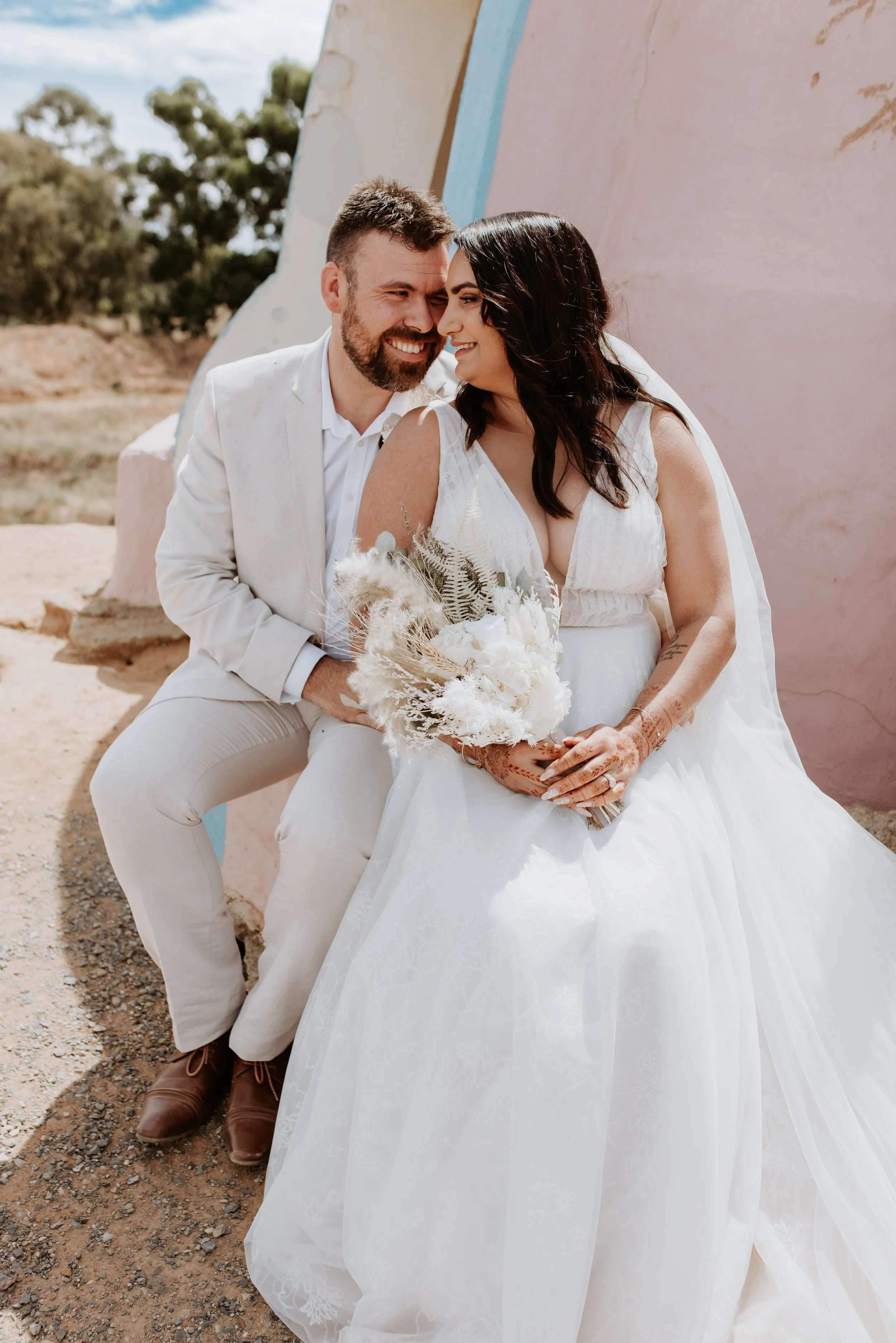 A couple, dressed in wedding attire, sitting close together outdoors, smiling at each other, with a pastel-colored wall and trees in the background.