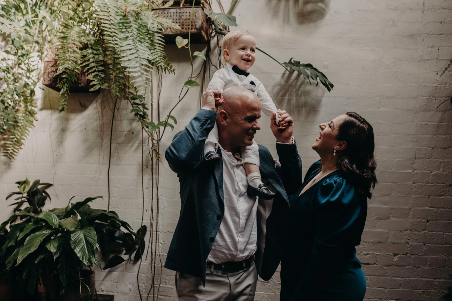A man, woman, and a young boy celebrating together indoors, with the man lifting the boy on his shoulders, all smiling and looking at each other, surrounded by plants and a white brick wall.