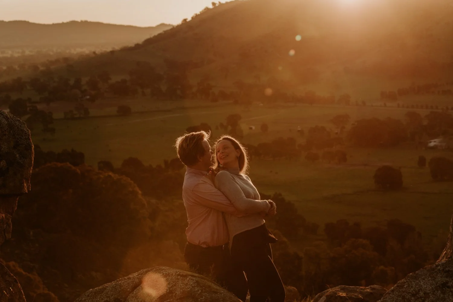 A couple embracing and smiling at sunset on a rocky hillside with a landscape of fields, trees, and hills in the background.