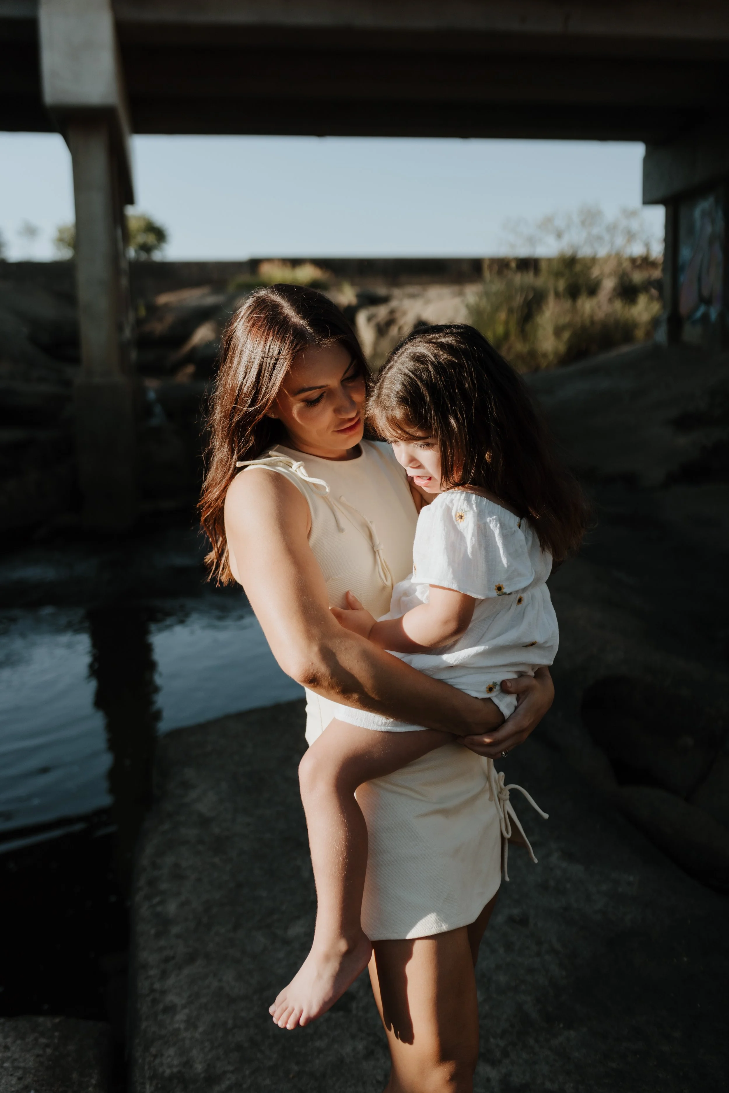 A woman holding a young girl in her arms under a bridge near water, both wearing light-colored clothing, during a sunny day.