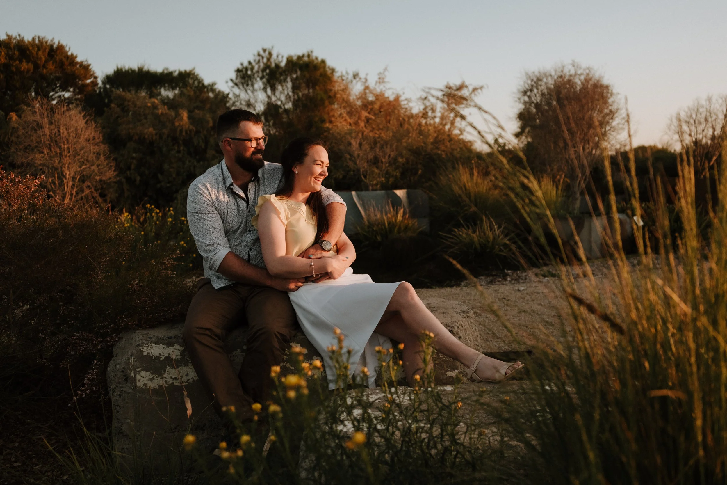 A happy couple, a man and a woman, sitting on a large rock outdoors during sunset with trees and plants in the background, smiling and enjoying each other's company.