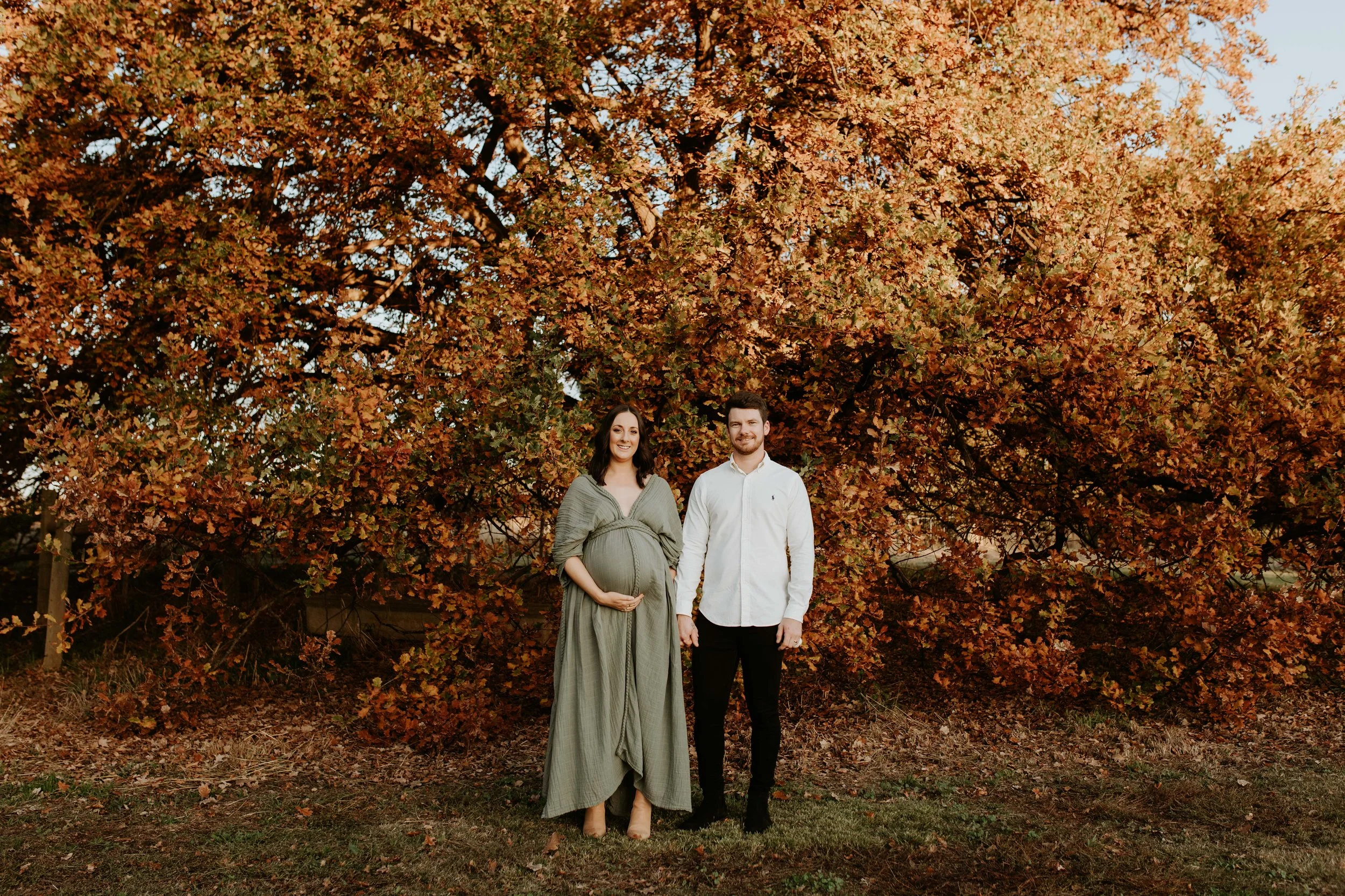 A couple holding hands, standing outdoors in front of a large, vibrant red and orange autumn tree, with the woman appearing to be pregnant.
