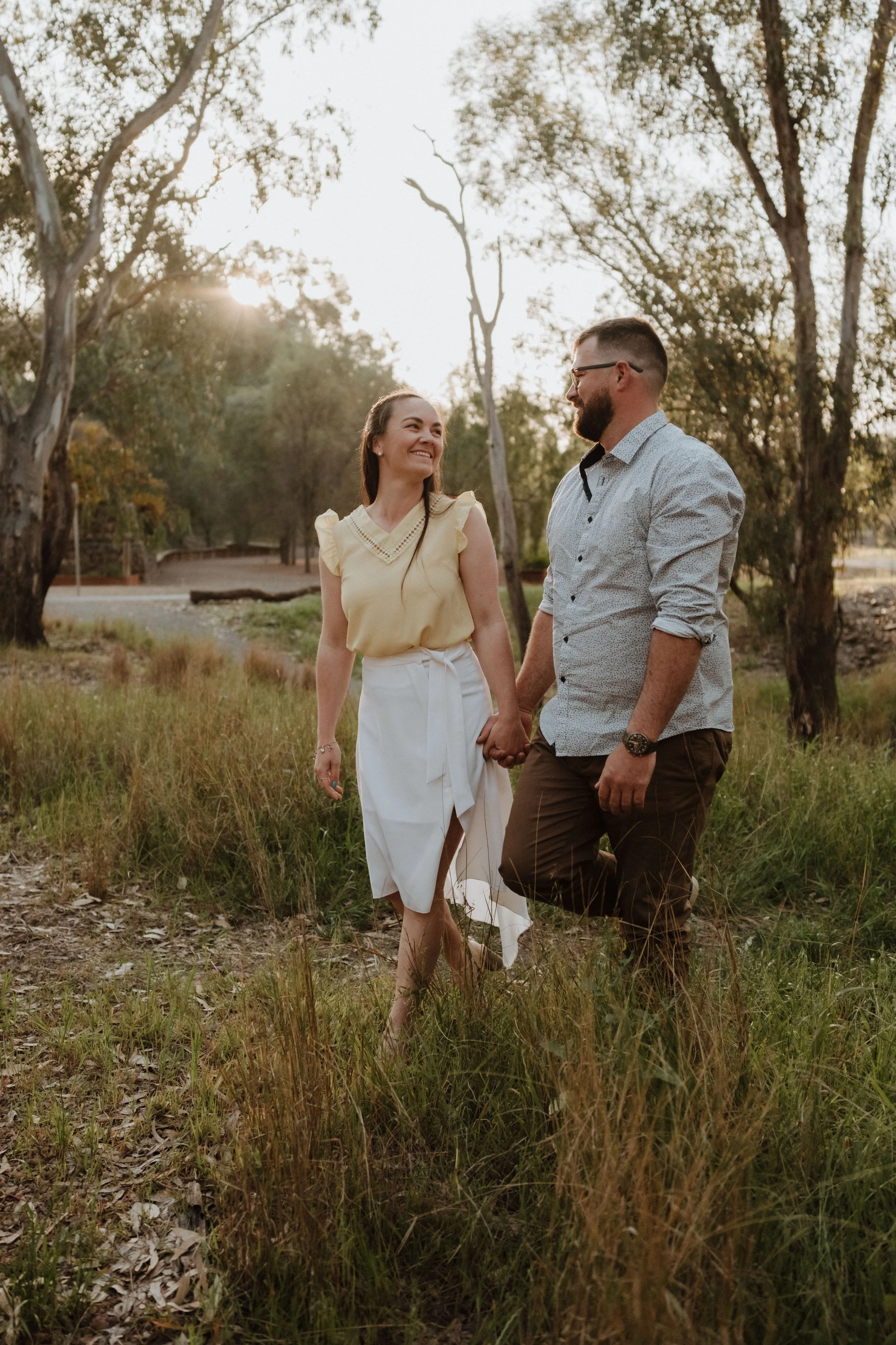 A couple holding hands and walking through a grassy field with trees in the background during sunset.