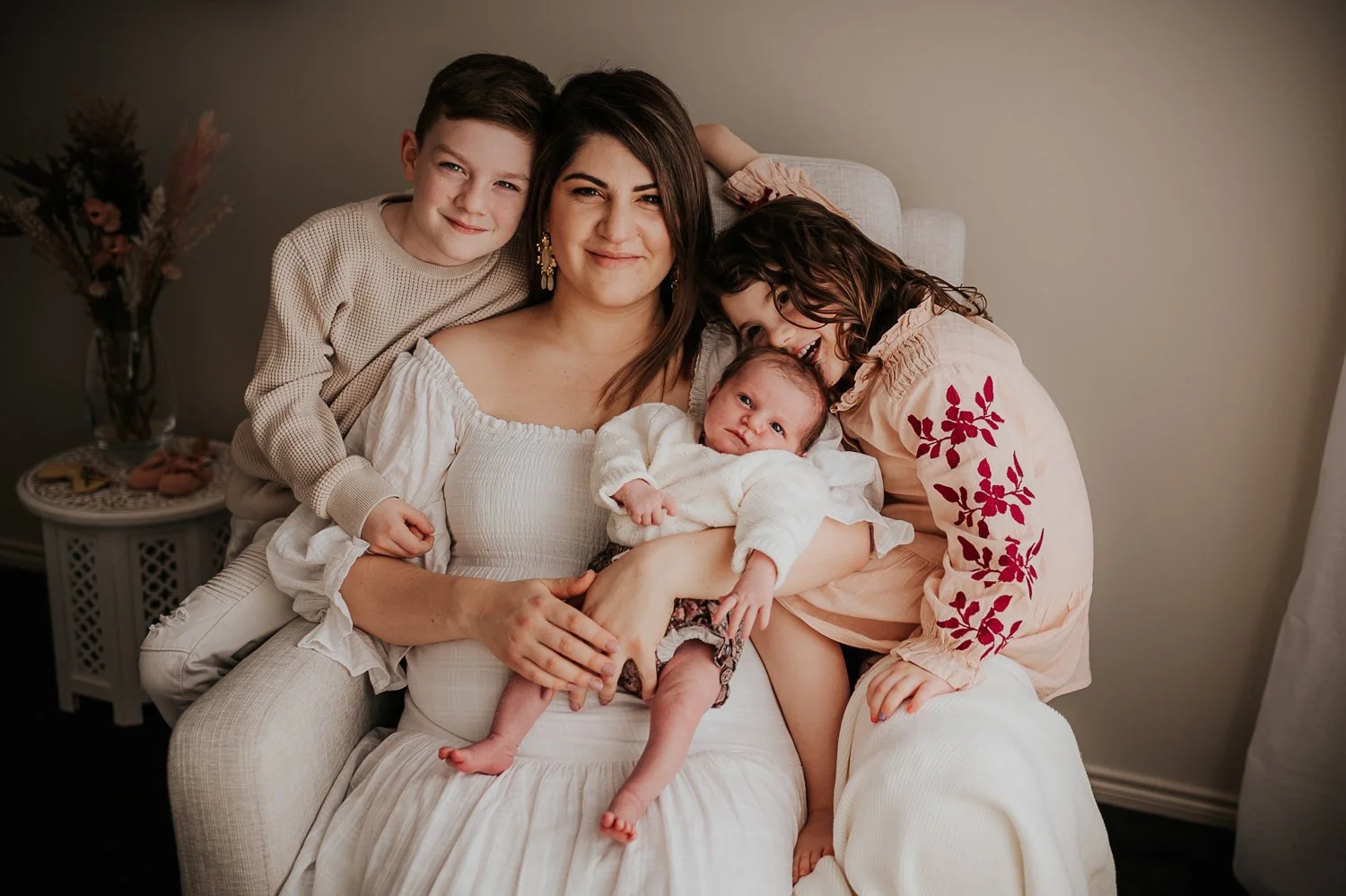 A woman sitting on a beige armchair with three children, one baby in her lap, two young girls leaning on her, all smiling and looking at the camera, in a cozy home setting.